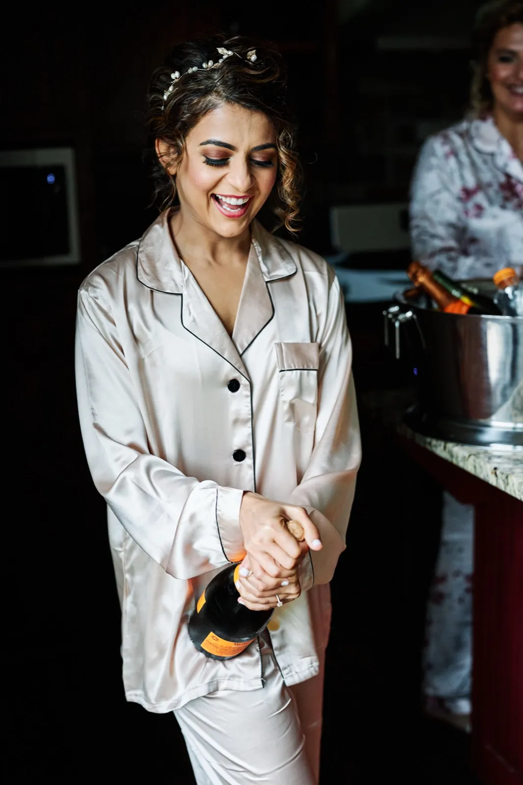 The bride laughs as she twists the cork from a champagne bottle in her satin pajamas, while another woman watches from beside an ice bucket in the background. — Tim Larsen Photography, Brainerd Lakes MN
