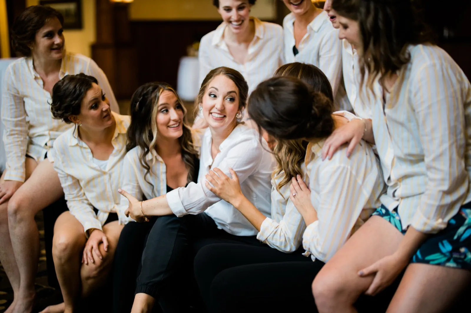 The bride laughs in the center of her bridesmaids, all wearing matching striped shirts, as they lean in around her in a getting-ready room. — Tim Larsen Photography, Brainerd Lakes MN