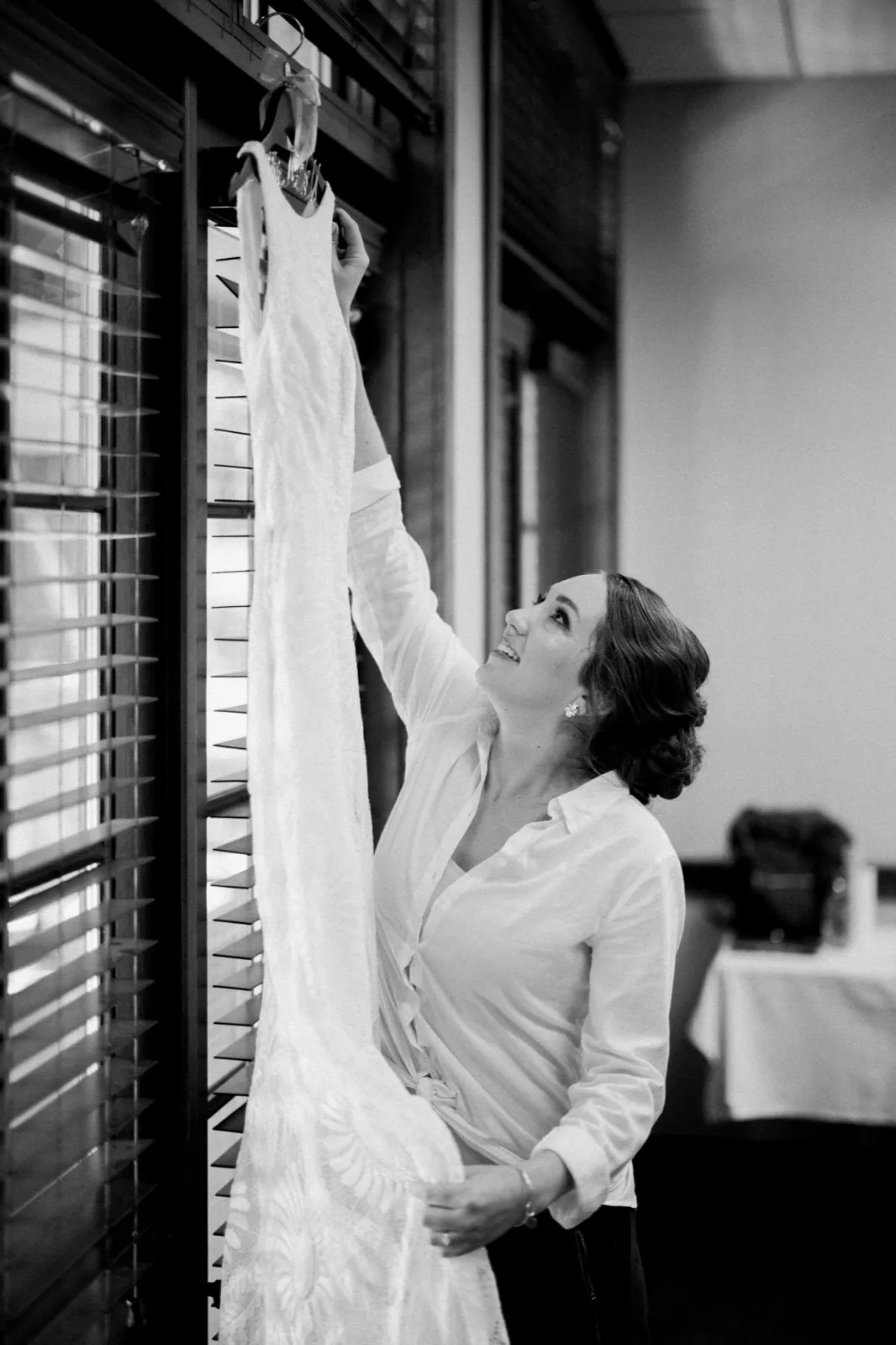 The bride reaches up to lift her wedding dress from a hanger by a window with horizontal blinds, smiling as she looks up at it. — Tim Larsen Photography, Brainerd Lakes MN