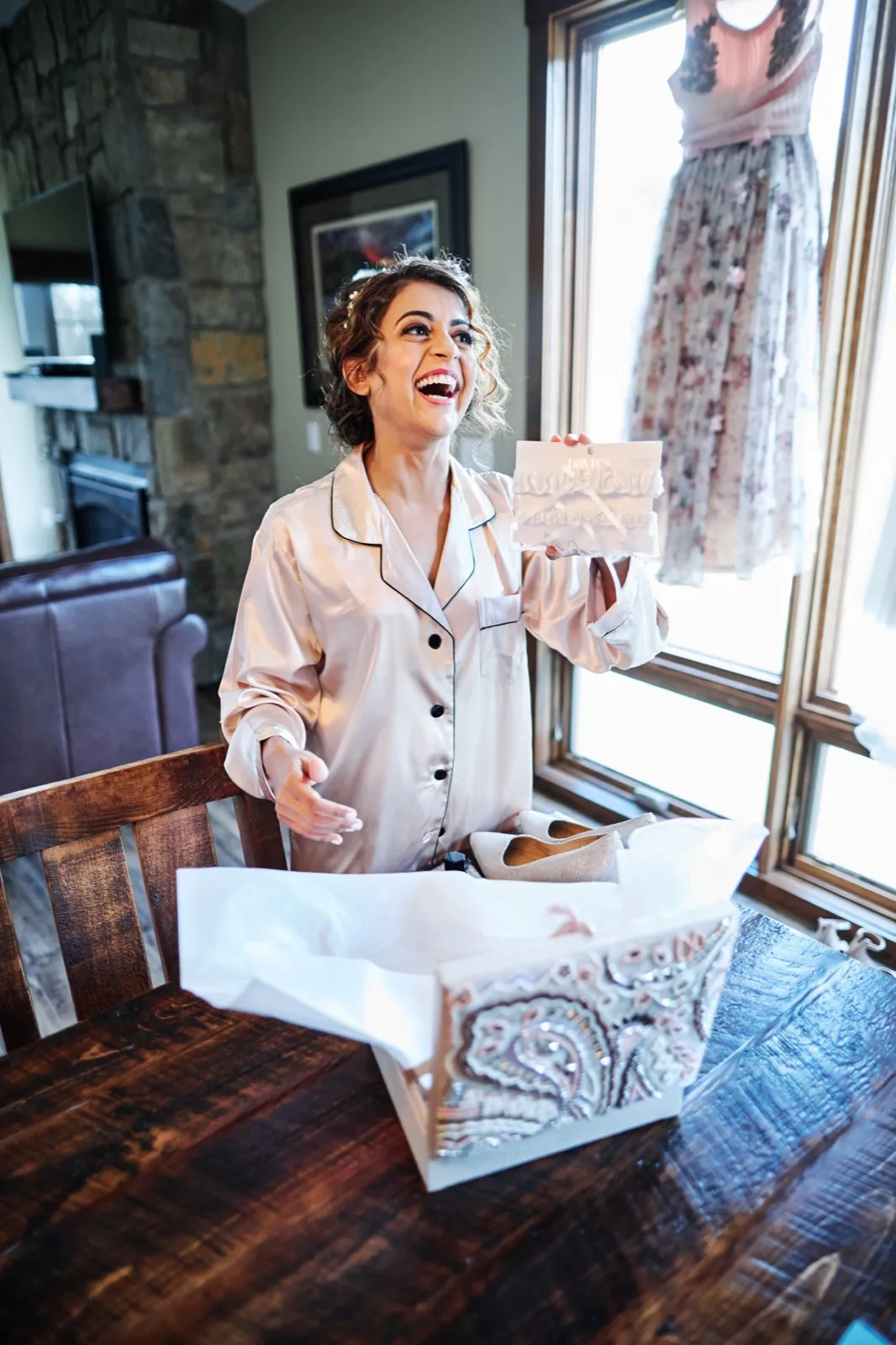 The bride in satin pajamas laughs openly while holding up a wedding garter, with her hanging dress and an opened shoe box visible nearby. — Tim Larsen Photography, Brainerd Lakes MN