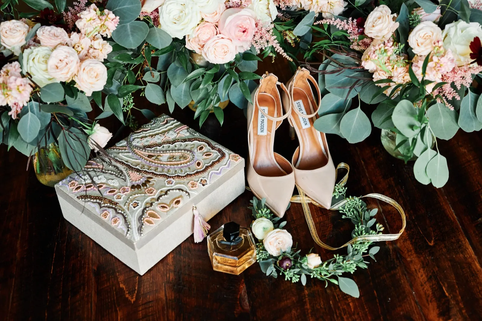 An overhead detail of nude heels, a perfume bottle, an embroidered paisley box, and a floral crown arranged among blush and ivory bouquets on a dark wood floor. — Tim Larsen Photography, Brainerd Lakes MN