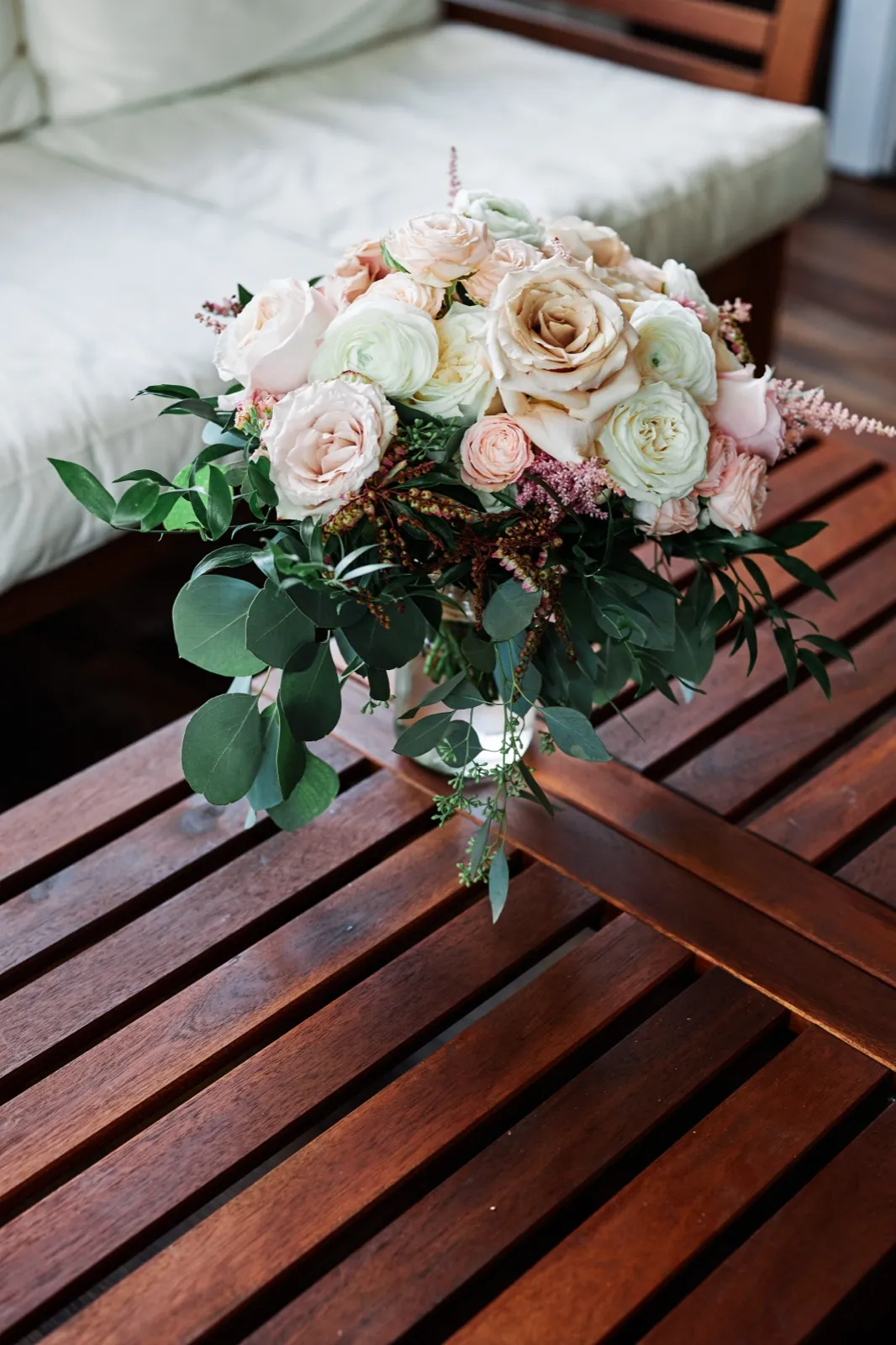 A bridal bouquet of blush roses, ivory ranunculus, and trailing eucalyptus rests on a dark slatted wood bench beside a cream cushioned chair. — Tim Larsen Photography, Brainerd Lakes MN
