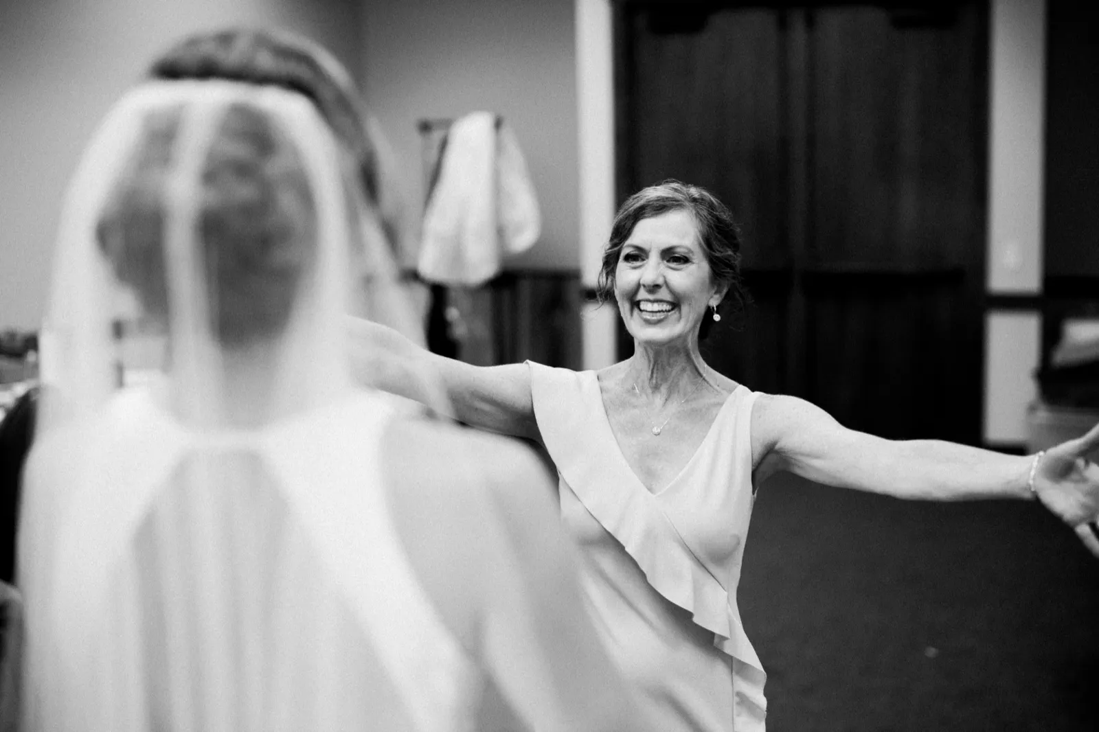 A mother stands with arms outstretched, beaming at her daughter, whose veiled figure is softly out of focus in the foreground of a bridal suite. — Tim Larsen Photography, Brainerd Lakes MN