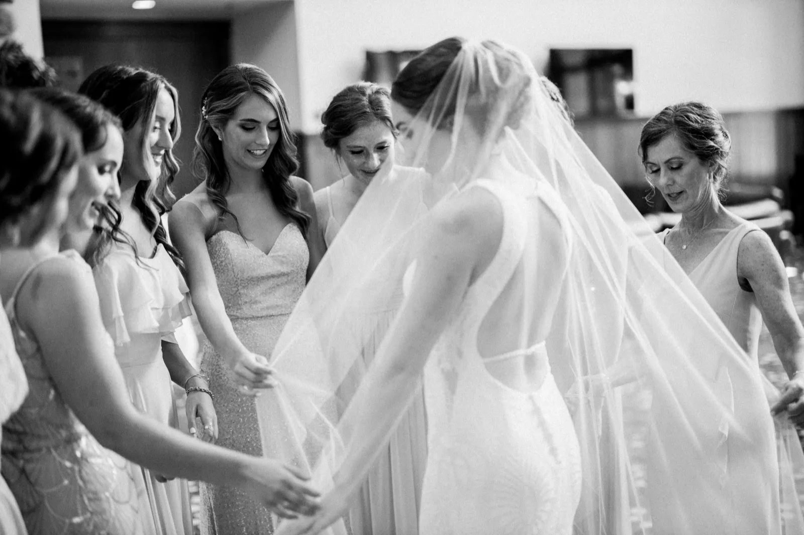 The bridesmaids stand in a circle around the bride, lifting and spreading her long veil and dress in a hotel suite, rendered in black and white. — Tim Larsen Photography, Brainerd Lakes MN