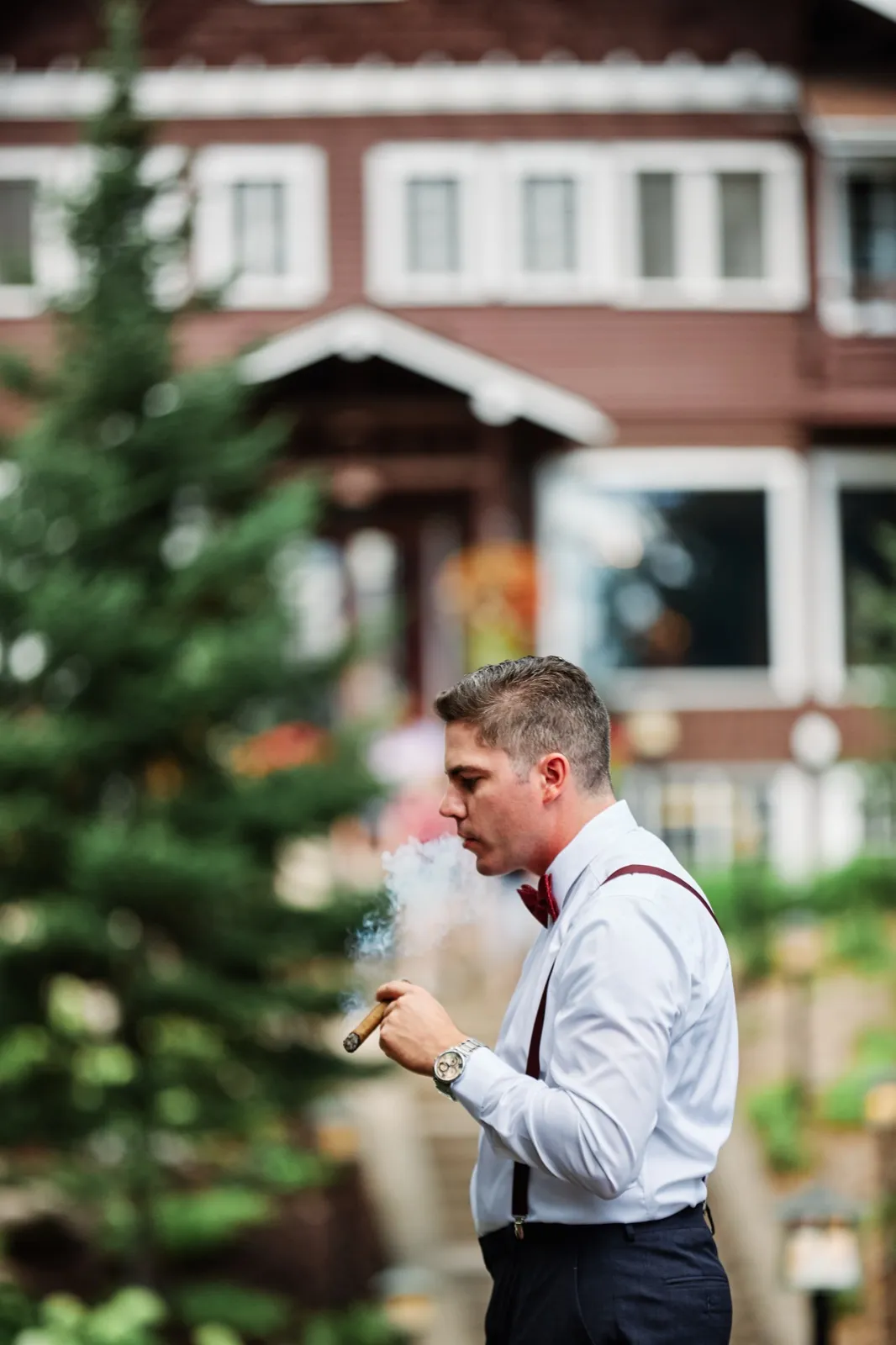 The groom in burgundy suspenders and bow tie exhales cigar smoke in profile outside the Grand View Lodge. — Tim Larsen Photography, Brainerd Lakes MN