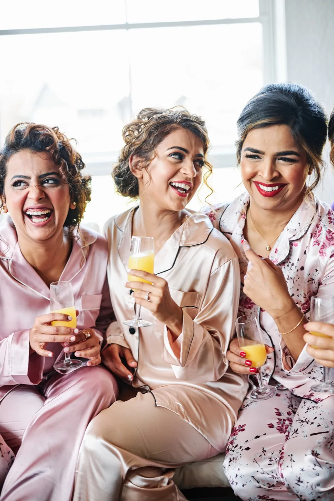 The bride and her bridesmaids laugh together in matching pink pajamas while holding mimosas in front of a bright window. — Tim Larsen Photography, Brainerd Lakes MN