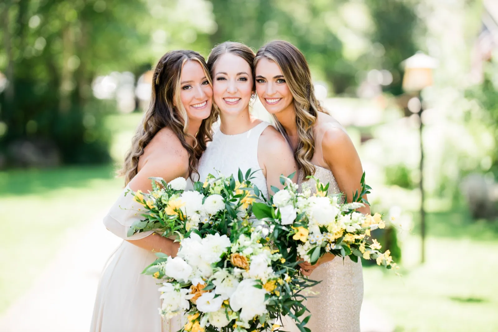 The bride stands between two bridesmaids, all three smiling at the camera with cheeks pressed close while holding large yellow and white bouquets on a sunlit garden path. — Tim Larsen Photography, Brainerd Lakes MN