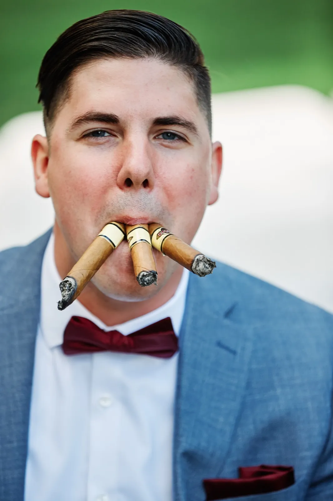 The groom in a light blue suit and burgundy bow tie stares directly at the camera with three lit cigars in his mouth. — Tim Larsen Photography, Brainerd Lakes MN
