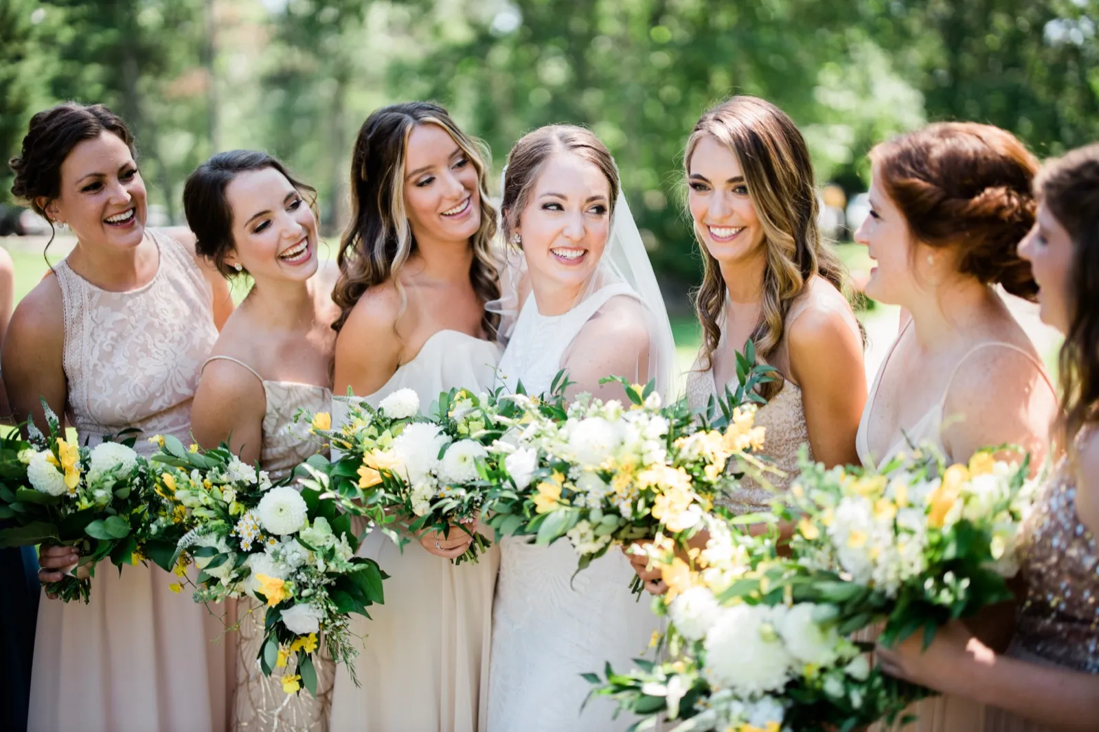 The bride laughs with her bridesmaids gathered close around her, all holding yellow and white bouquets on a sunlit lawn. — Tim Larsen Photography, Brainerd Lakes MN