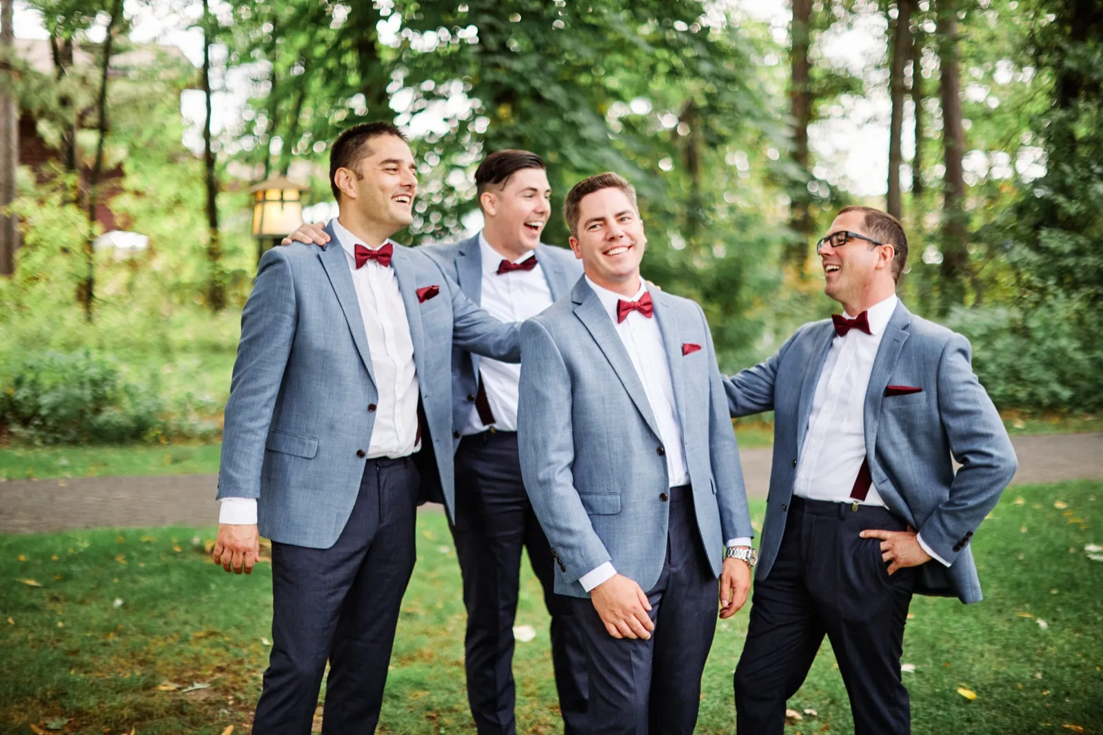 The groom and three groomsmen in dusty-blue suits with burgundy bow ties laugh together on a wooded path. — Tim Larsen Photography, Brainerd Lakes MN