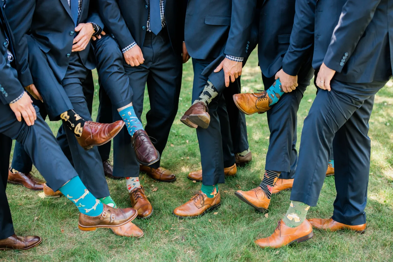 Seven groomsmen in navy suits lift their pant cuffs to reveal mismatched patterned socks above polished dress shoes on the grass. — Tim Larsen Photography, Brainerd Lakes MN