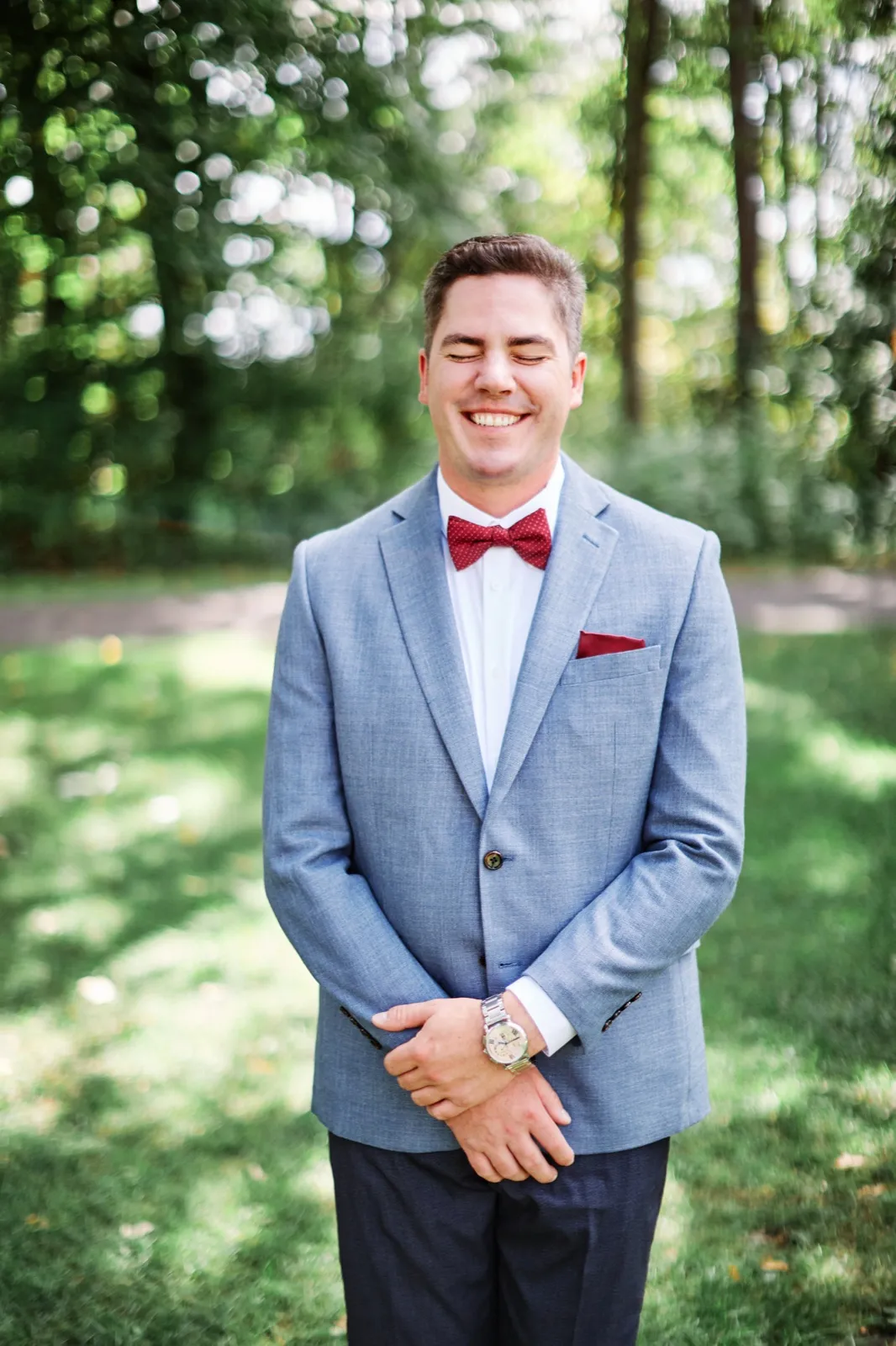 The groom laughs with his eyes closed, wearing a light blue jacket and red bow tie, standing on a sunlit lawn with blurred trees behind him. — Tim Larsen Photography, Brainerd Lakes MN