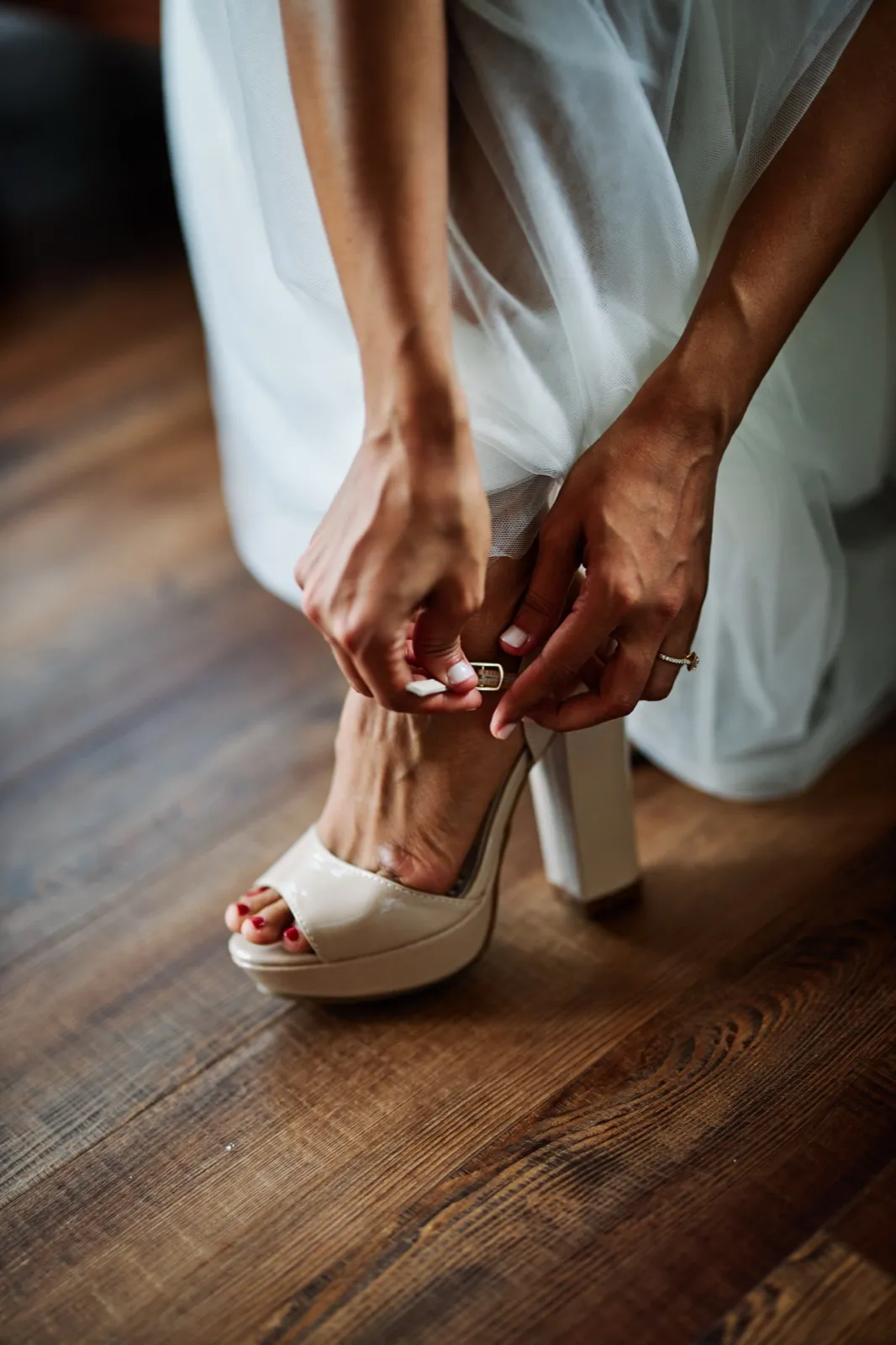The bride bends down to fasten the buckle on a nude platform heel, her engagement ring visible on her hand against the wood floor. — Tim Larsen Photography, Brainerd Lakes MN