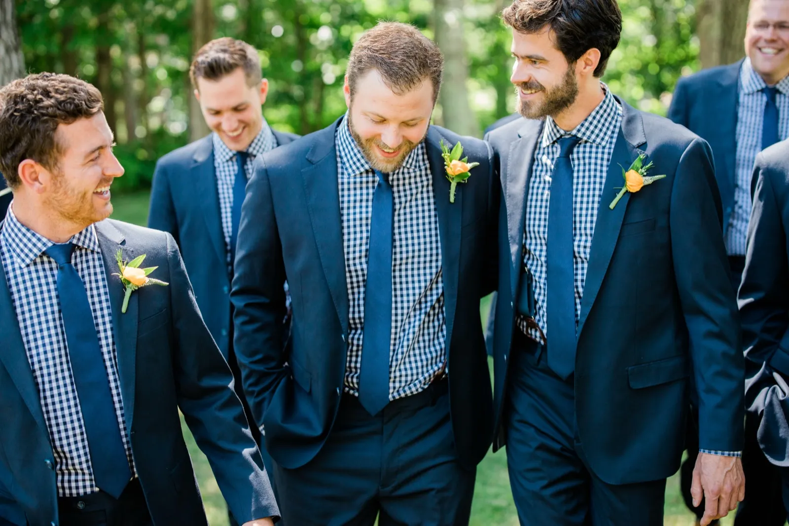 The groom looks down laughing as his groomsmen in navy suits and gingham shirts walk alongside him through a wooded area. — Tim Larsen Photography, Brainerd Lakes MN