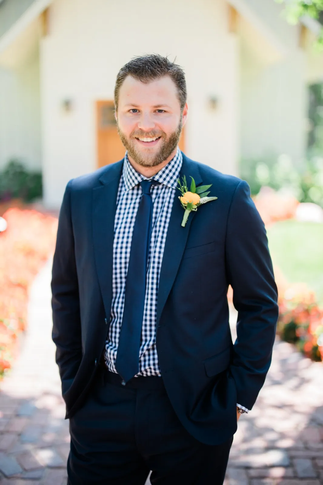 The groom stands smiling in a navy suit and gingham shirt on a brick walkway outside the Grand View Lodge chapel, with red flowerbeds blurred on either side. — Tim Larsen Photography, Brainerd Lakes MN