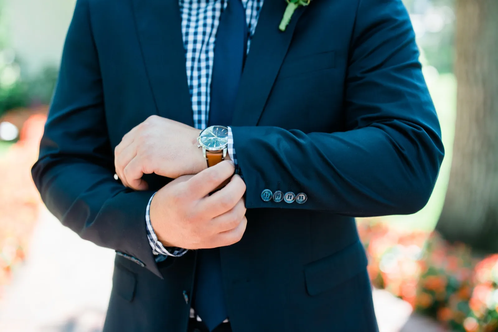 The groom adjusts his leather-strapped watch over the cuff of a navy suit, with a gingham shirt and boutonniere visible at his lapel. — Tim Larsen Photography, Brainerd Lakes MN