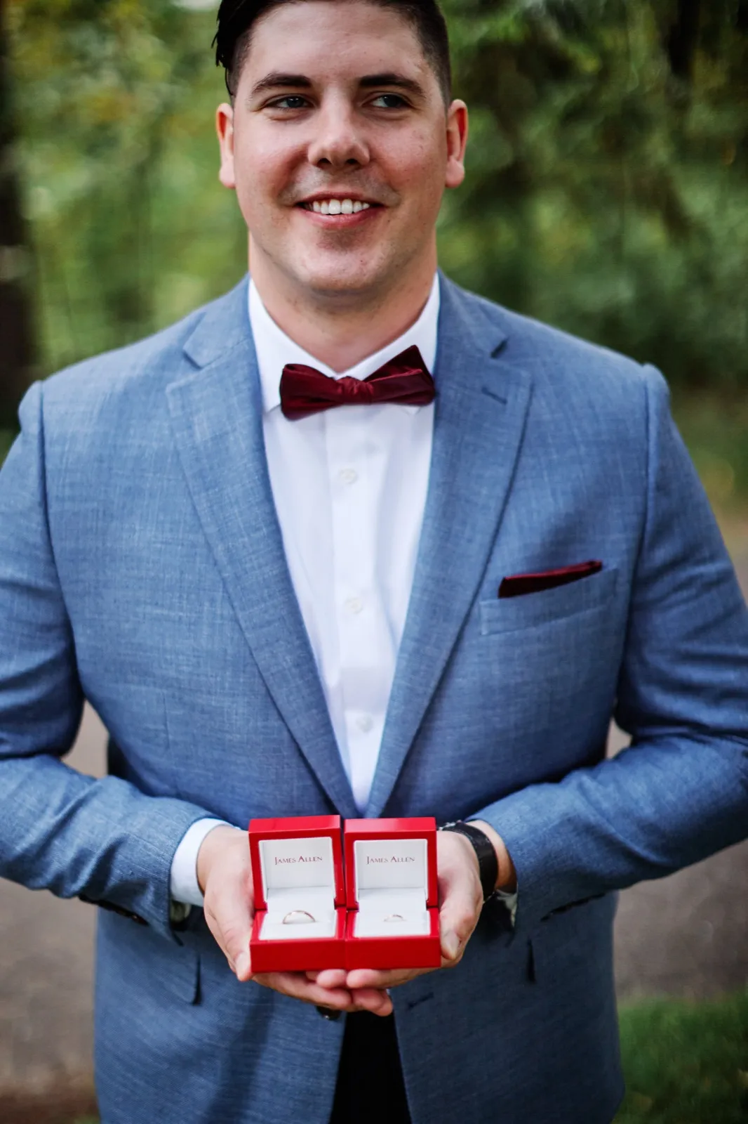 The groom in a blue suit and burgundy bow tie smiles at the camera while holding two open red James Allen ring boxes displaying the wedding bands. — Tim Larsen Photography, Brainerd Lakes MN