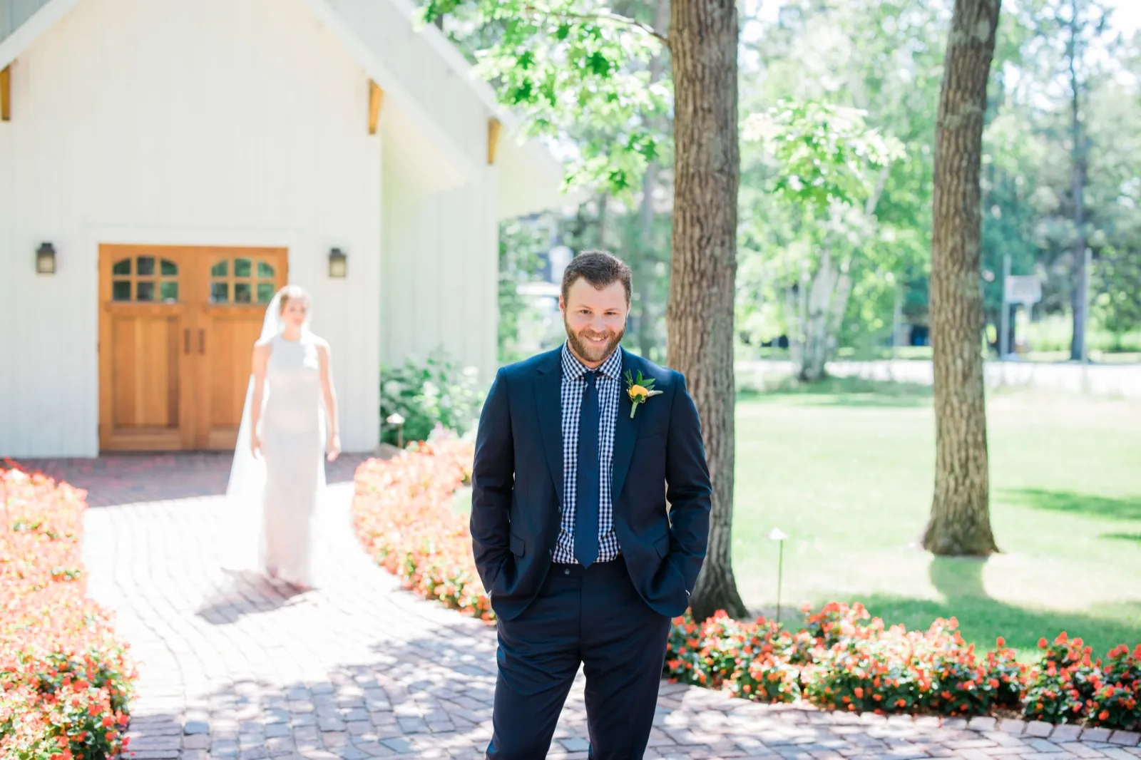 The groom waits on a brick path lined with orange flowers as the bride approaches softly out of focus from the white chapel doors behind him. — Tim Larsen Photography, Brainerd Lakes MN