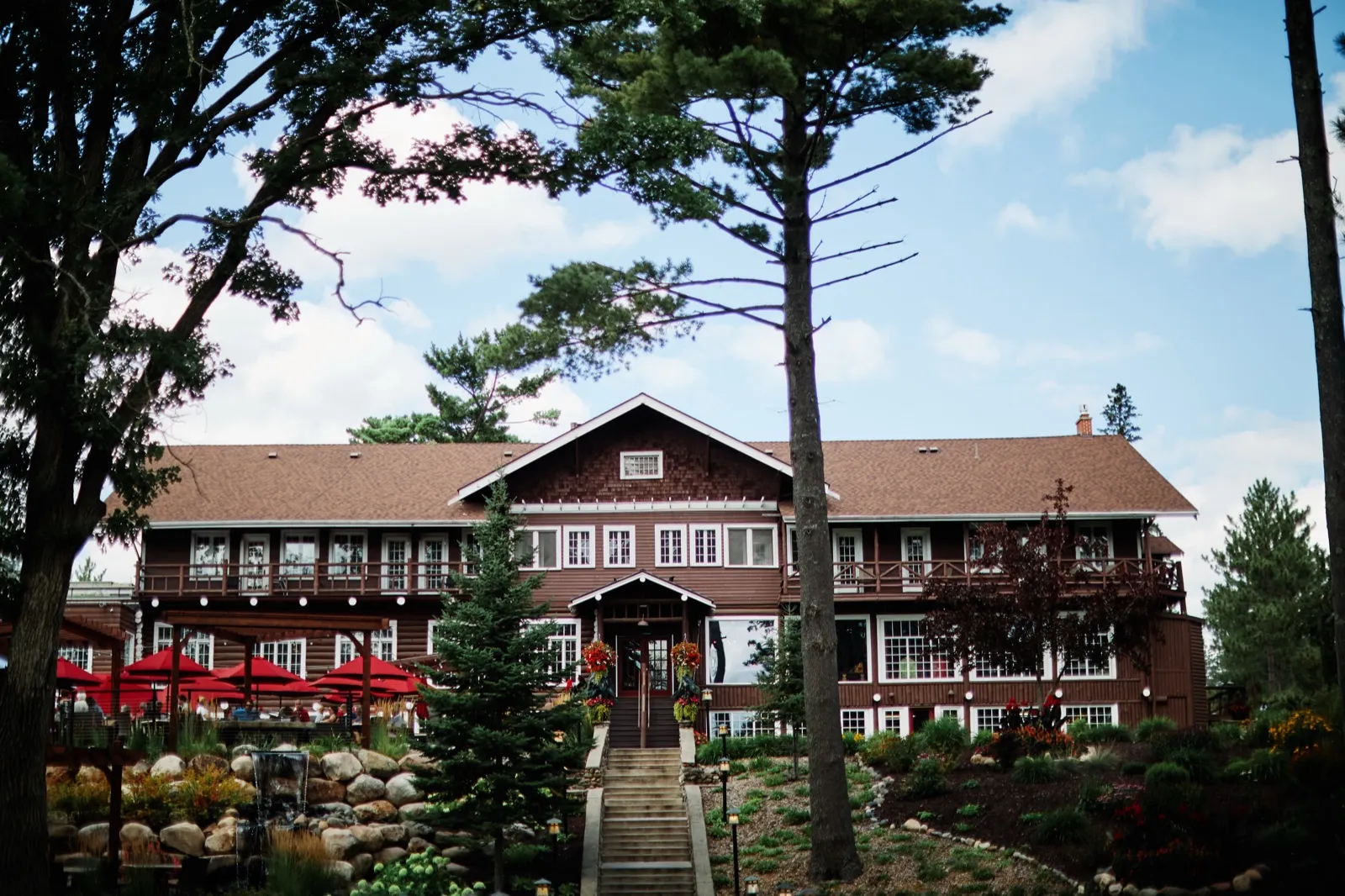 The historic Grand View Lodge main building stands behind tall pines, with stone steps leading up to its covered entrance and red patio umbrellas visible to the side. — Tim Larsen Photography, Brainerd Lakes MN