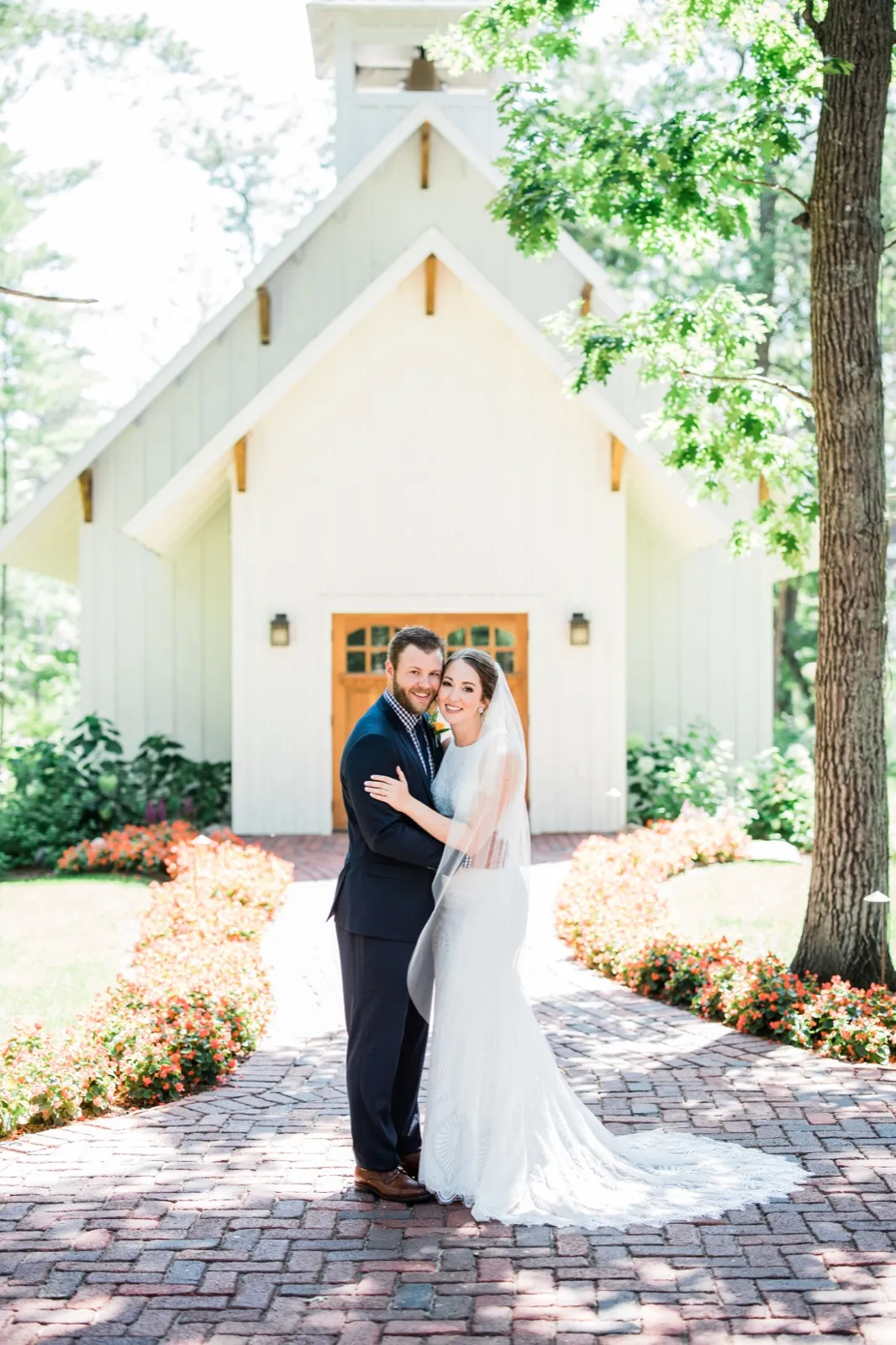 The bride and groom embrace on a brick walkway in front of a white chapel at Grand View Lodge, her long veil and lace train trailing behind her. — Tim Larsen Photography, Brainerd Lakes MN
