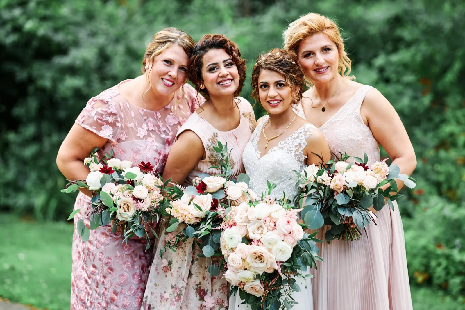 The bride stands with three bridesmaids in blush and floral dresses, all holding lush bouquets of pale roses and greenery against a leafy green background. — Tim Larsen Photography, Brainerd Lakes MN