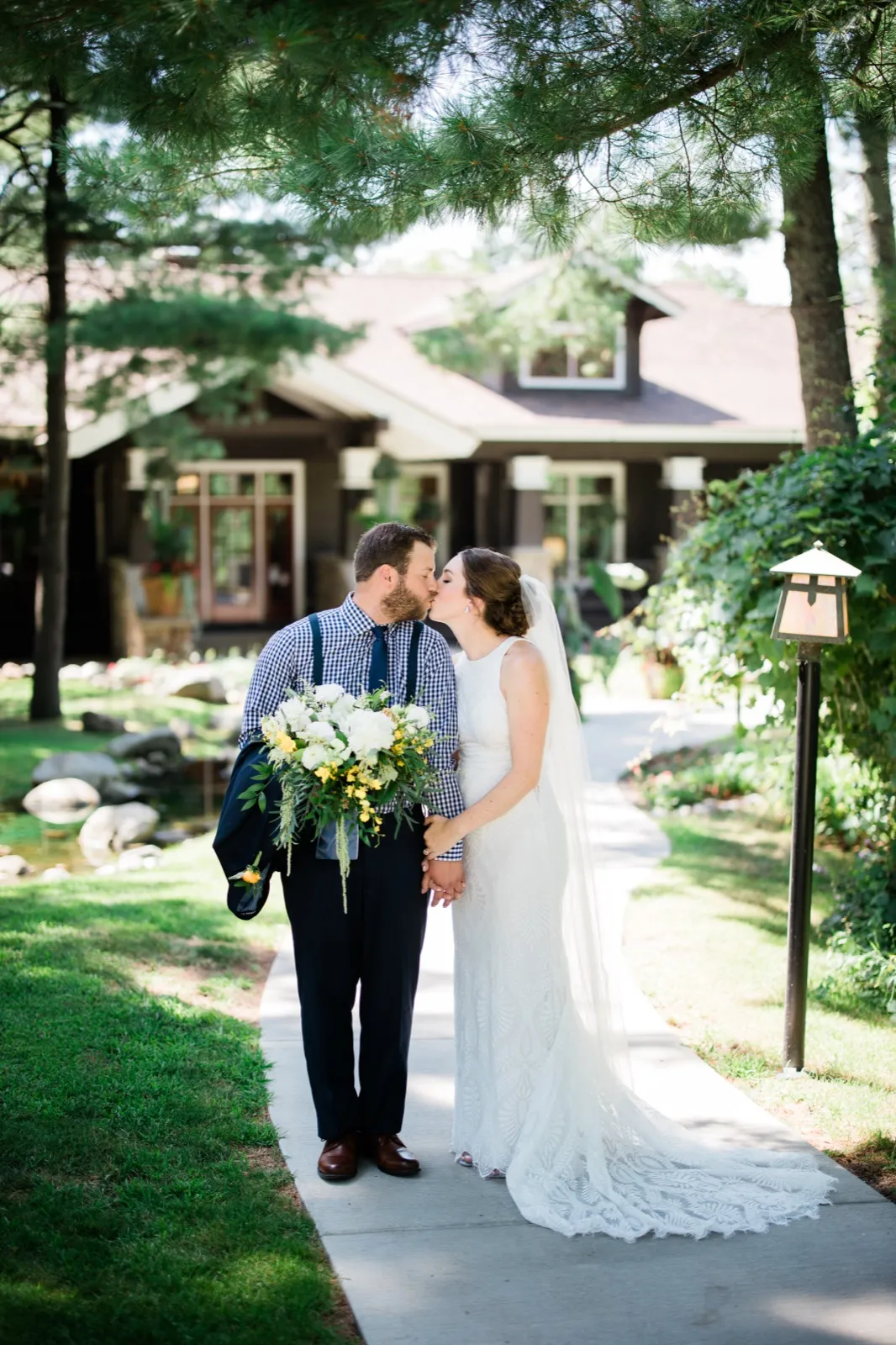 The bride and groom share a kiss on a sunlit walkway with the Grand View Lodge craftsman exterior and a lantern behind them, her long lace train trailing on the pavement. — Tim Larsen Photography, Brainerd Lakes MN
