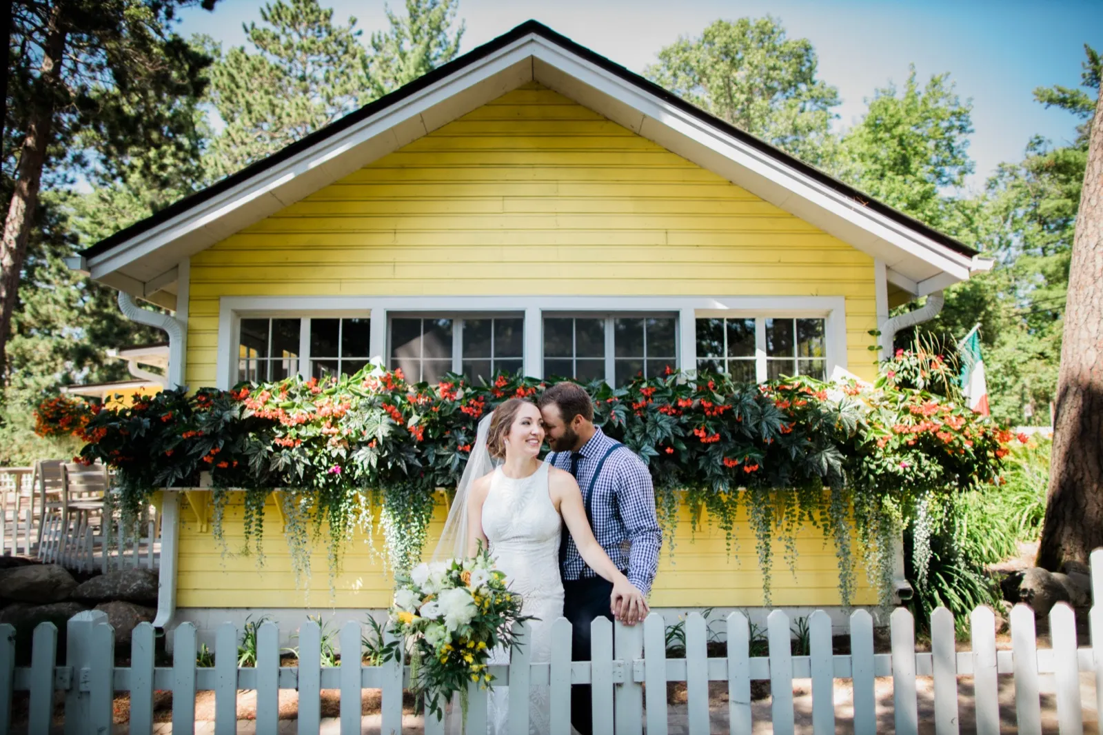 The bride and groom embrace behind a white picket fence in front of a yellow cottage with flower boxes overflowing with red and green blooms at Grand View Lodge. — Tim Larsen Photography, Brainerd Lakes MN