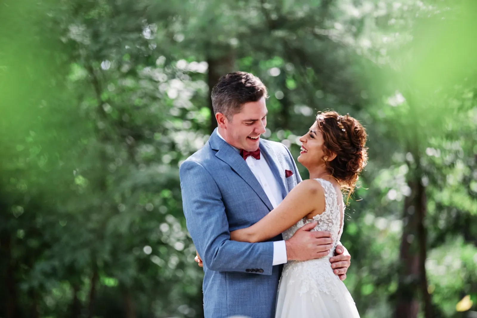 The bride laughs up at the groom as they embrace among backlit summer trees, her lace dress and his blue suit catching warm afternoon light. — Tim Larsen Photography, Brainerd Lakes MN