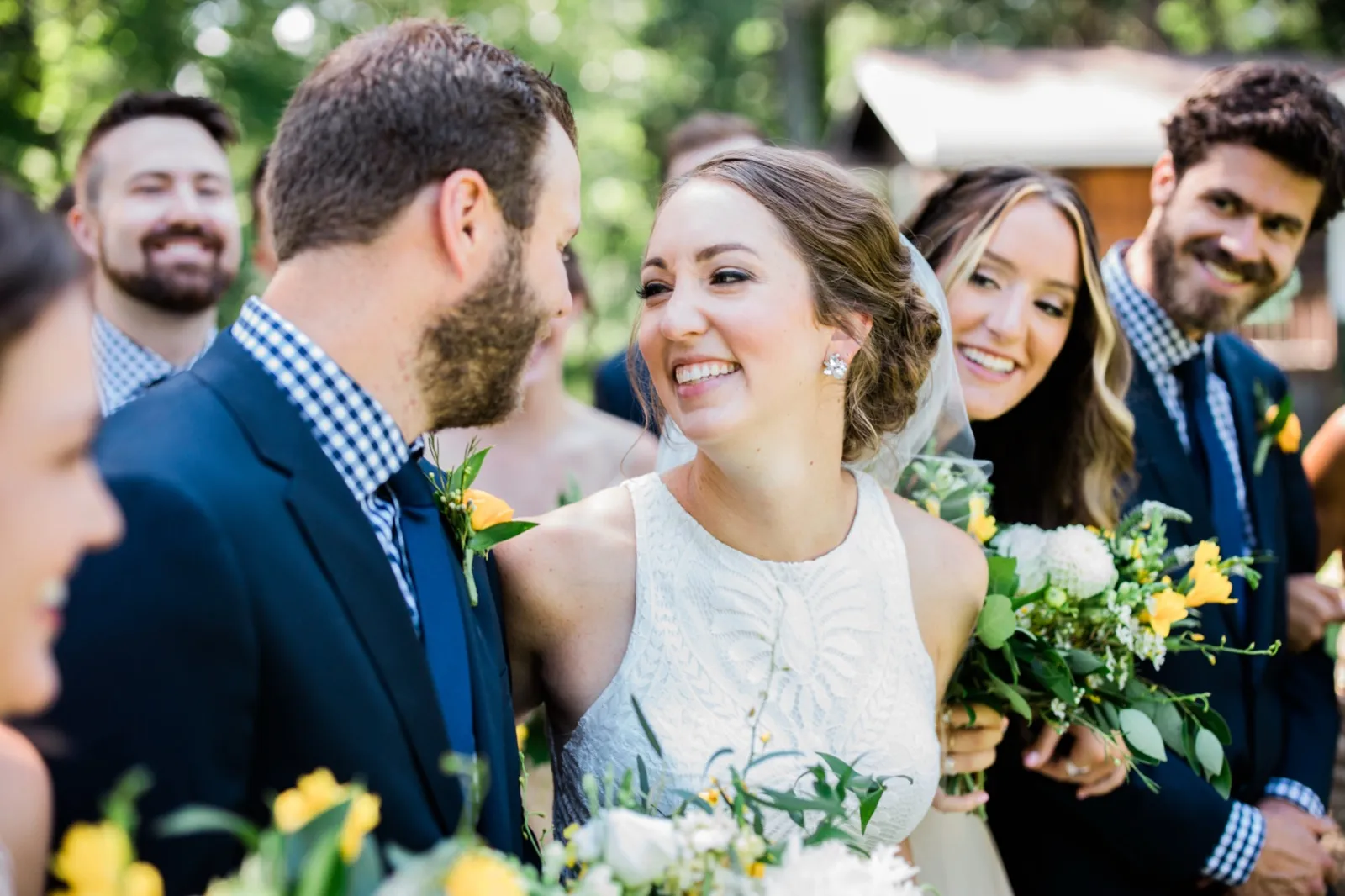 The bride smiles up at the groom while a bridesmaid laughs warmly beside her, surrounded by the bridal party in dappled outdoor light. — Tim Larsen Photography, Brainerd Lakes MN