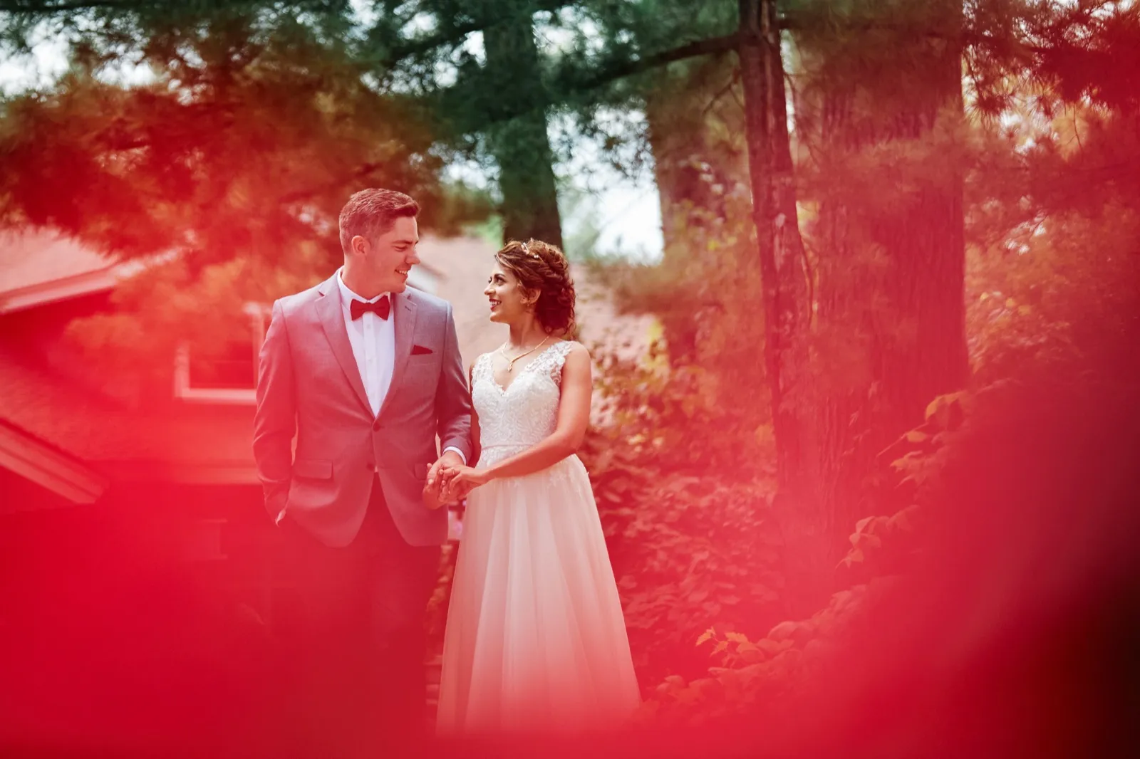 The bride and groom hold hands and smile at each other on a wooded path, framed by out-of-focus red foliage in the foreground. — Tim Larsen Photography, Brainerd Lakes MN