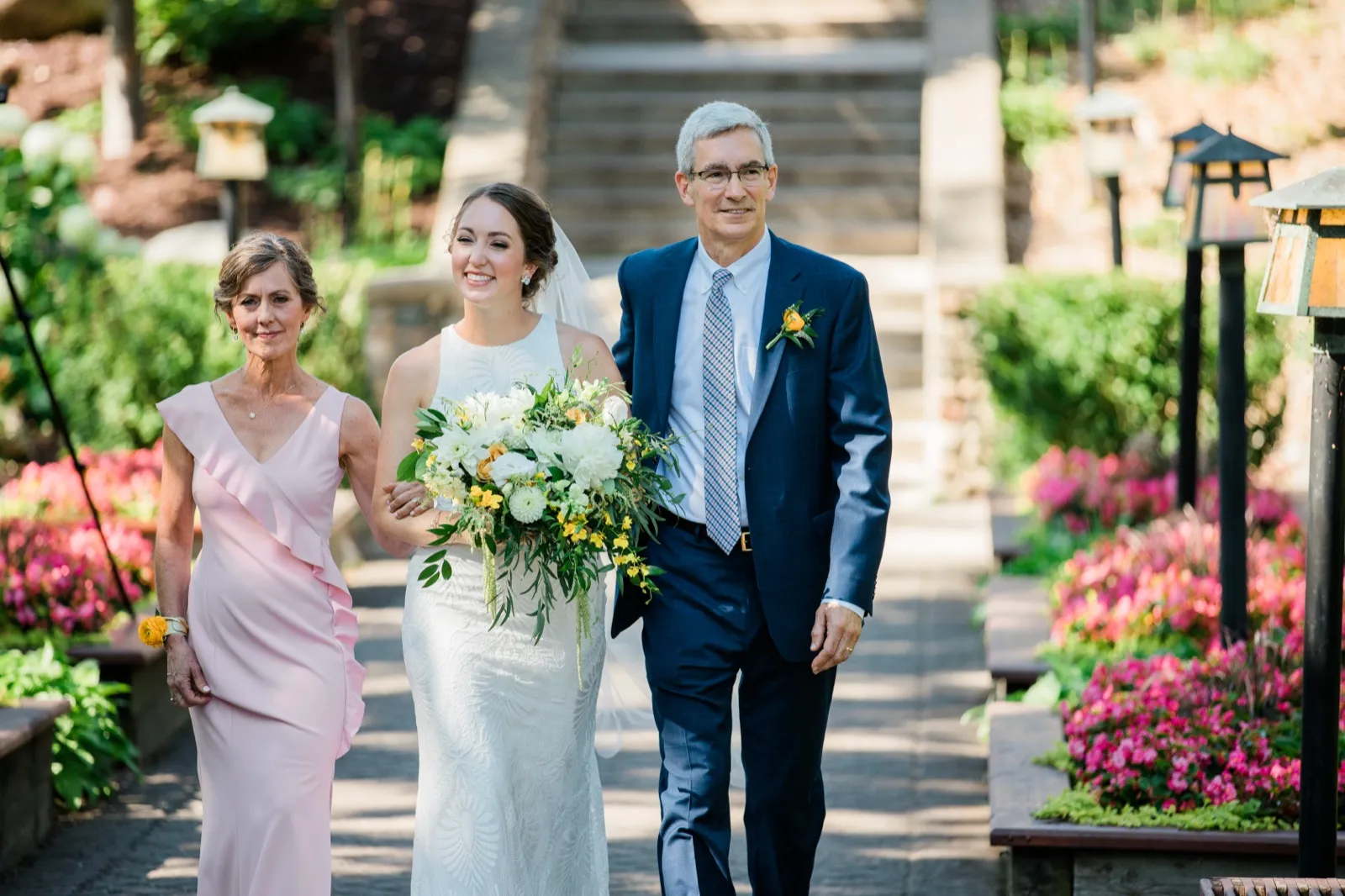 The bride walks down a flower-lined garden path between her parents, smiling and holding a large bouquet, with Grand View Lodge's stone staircase rising behind them. — Tim Larsen Photography, Brainerd Lakes MN