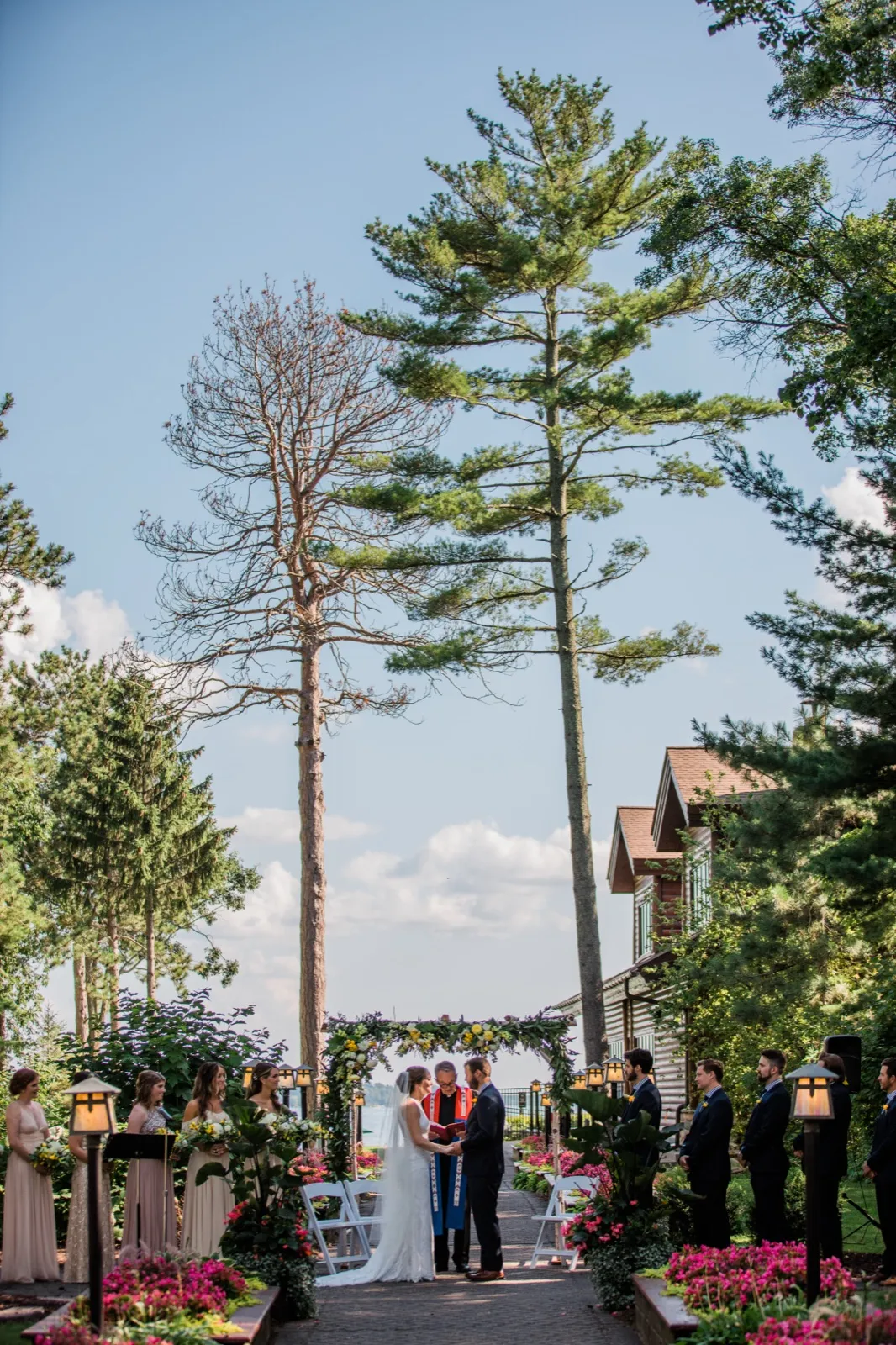 A wide view of an outdoor ceremony at Grand View Lodge shows the couple and officiant at a floral arch framed by towering pines, with the bridal party flanking the garden aisle and the lake visible beyond. — Tim Larsen Photography, Brainerd Lakes MN