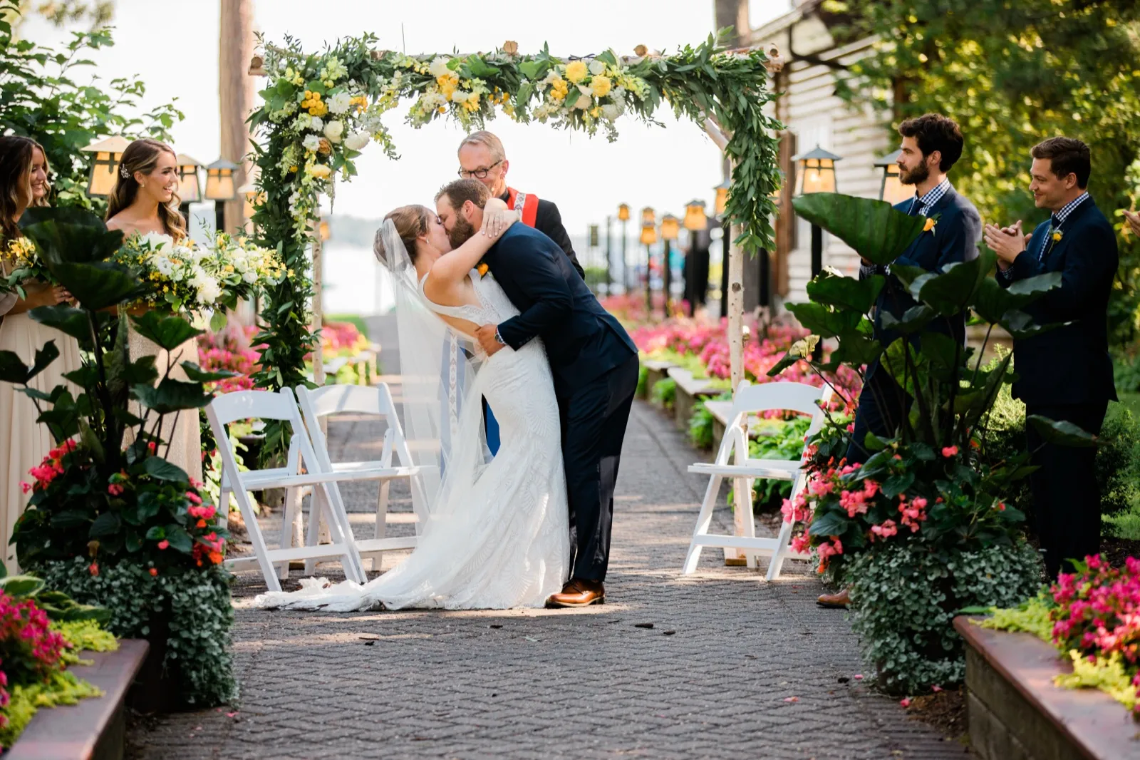 The bride and groom share their first kiss beneath a floral arch on a lakeside brick aisle while their bridal party watches and applauds. — Tim Larsen Photography, Brainerd Lakes MN
