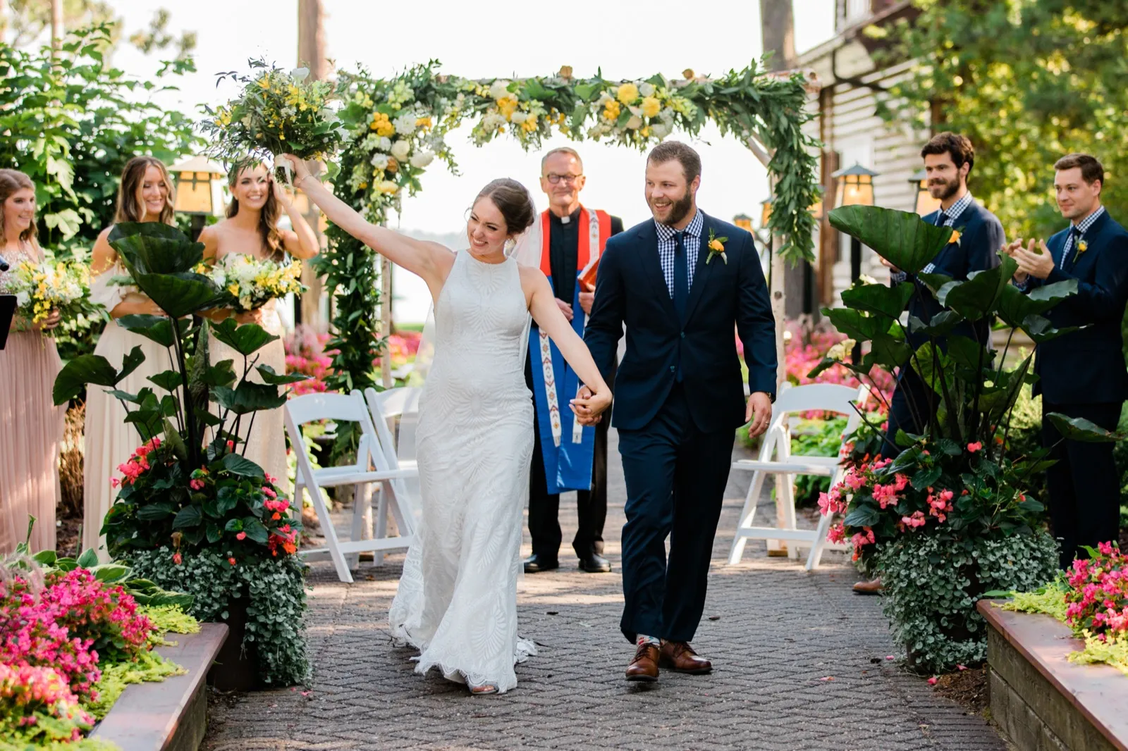 The bride raises her bouquet overhead in celebration as she walks down the aisle hand-in-hand with the groom, framed by a floral arch and clapping bridal party. — Tim Larsen Photography, Brainerd Lakes MN