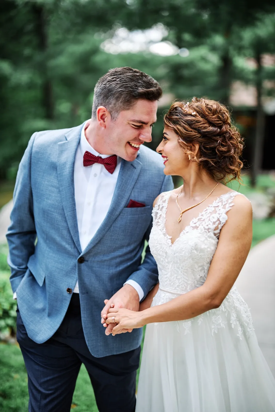 The bride and groom hold hands and laugh while looking at each other on a wooded pathway, her lace gown and his blue suit with burgundy bow tie catching soft daylight. — Tim Larsen Photography, Brainerd Lakes MN
