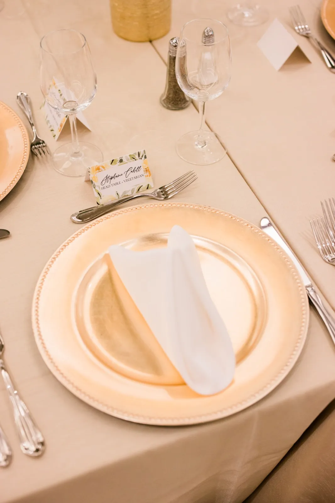 An overhead view of a head-table place setting with a gold charger plate, white folded napkin, floral place card reading Stephanie Cahill, and stemware on a champagne-colored linen. — Tim Larsen Photography, Brainerd Lakes MN