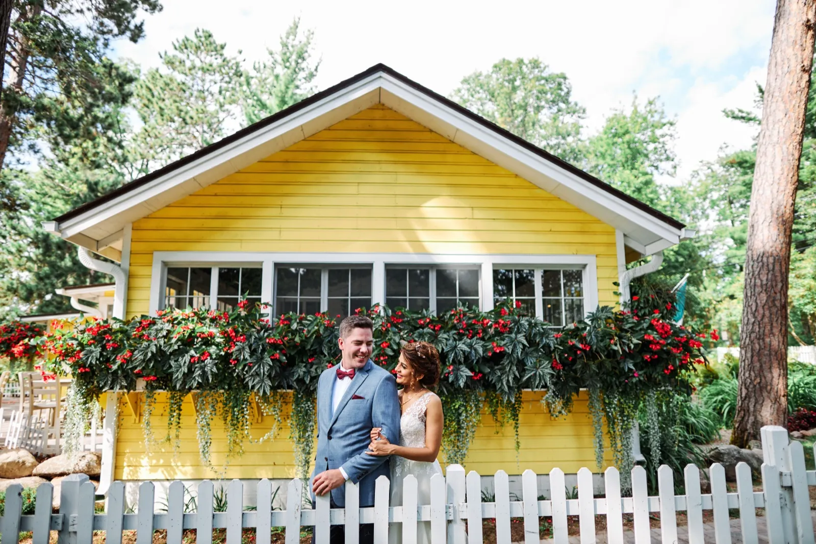 The couple stands close together in front of a yellow cottage with red and green flower-filled window boxes behind a white picket fence at Grand View Lodge. — Tim Larsen Photography, Brainerd Lakes MN