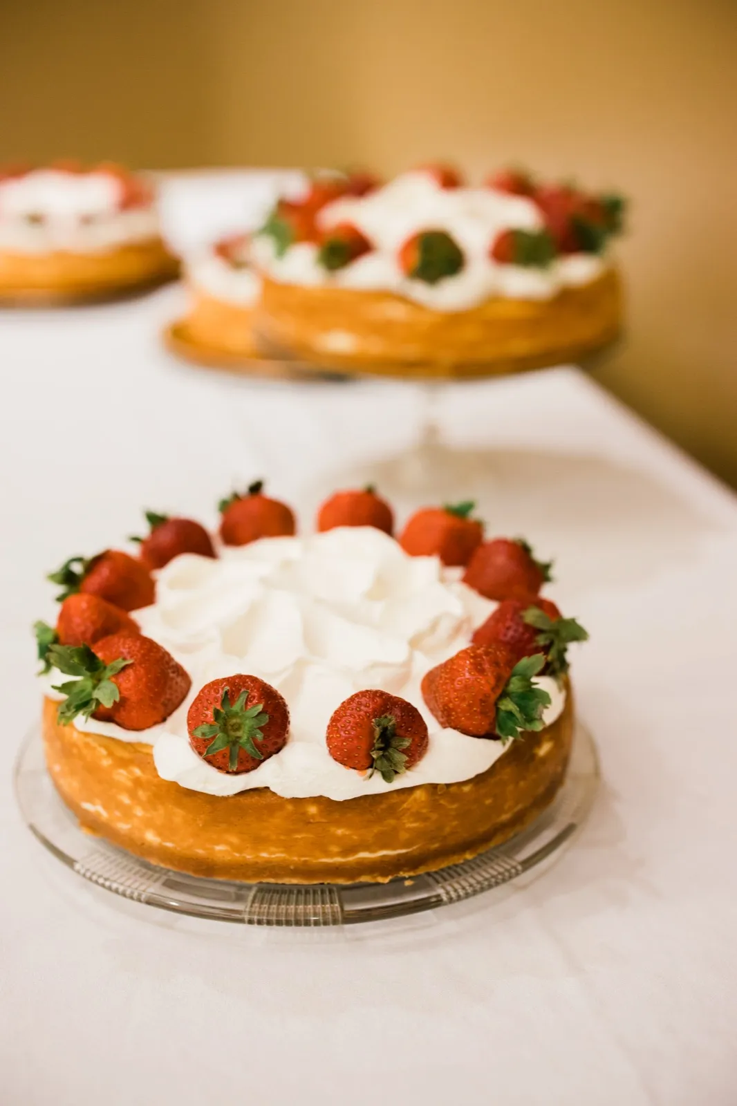 Two strawberry-topped cheesecakes with whipped cream sit on glass stands atop a white linen-covered table. — Tim Larsen Photography, Brainerd Lakes MN
