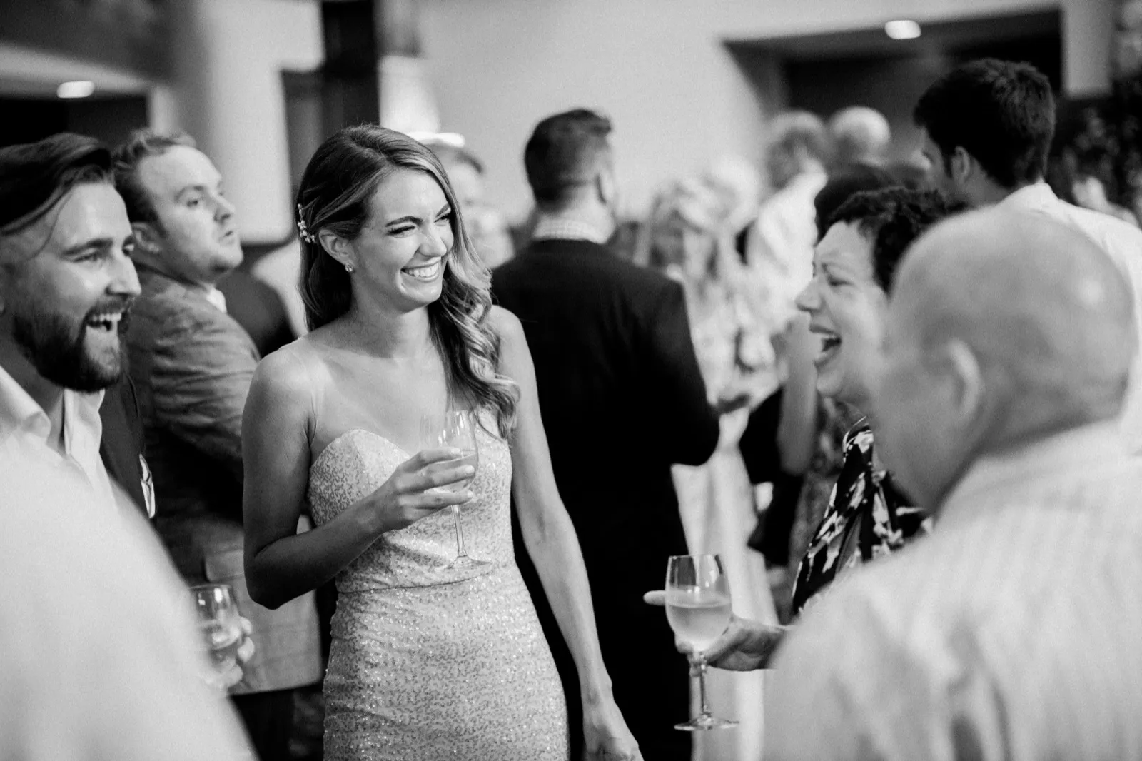 The bride laughs brightly while holding a champagne flute, surrounded by guests during cocktail hour in a black-and-white frame. — Tim Larsen Photography, Brainerd Lakes MN