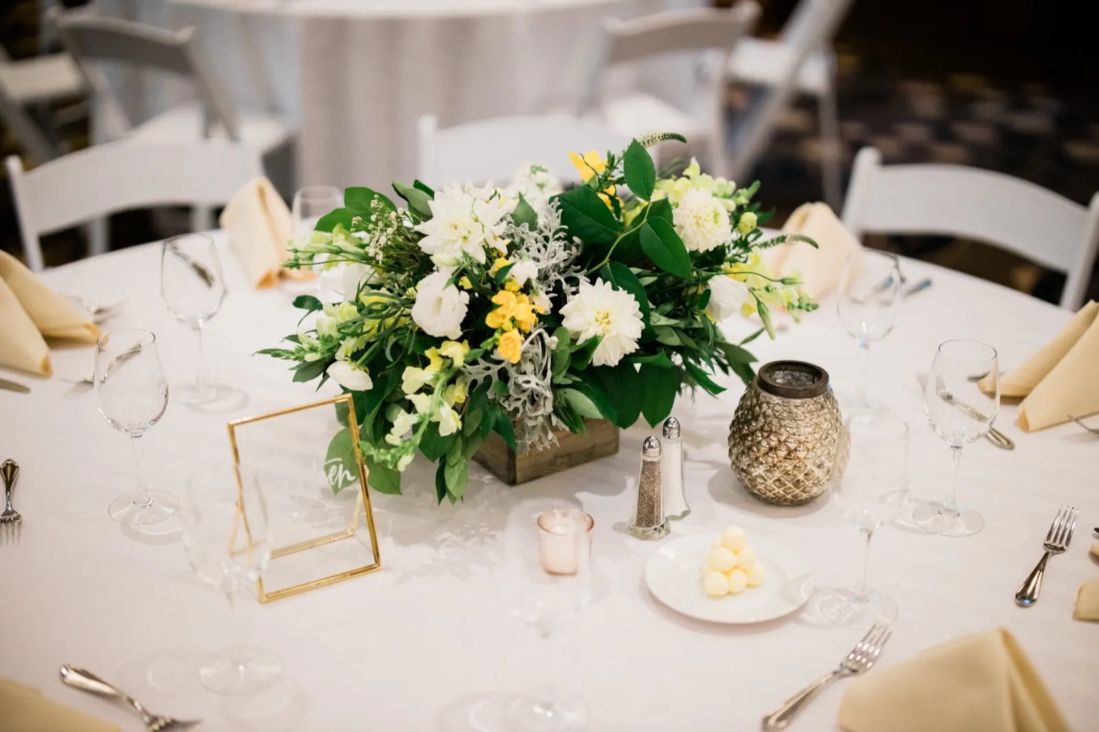 A white and yellow floral centerpiece in a wooden box sits at the center of a round reception table set with wine glasses, pale napkins, a gold place-card frame, and a textured silver votive. — Tim Larsen Photography, Brainerd Lakes MN