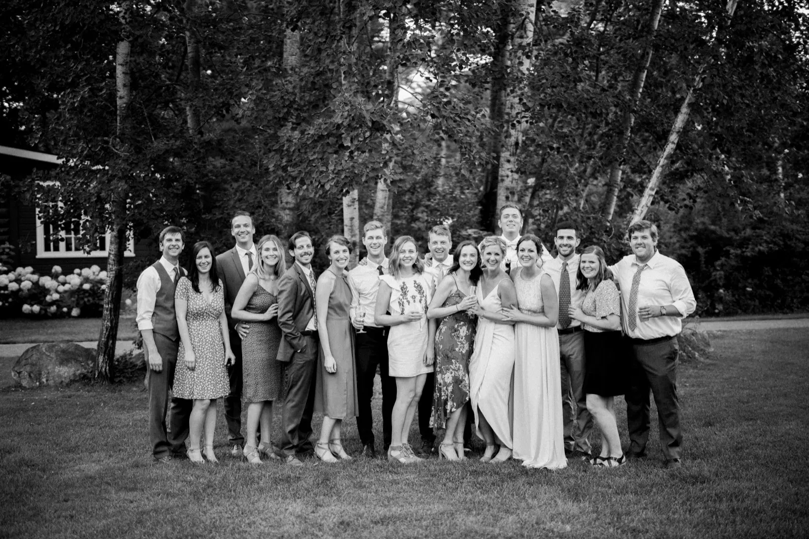 A large group of wedding guests including the bride and groom stand together on a lawn outside a wooded cabin at Grand View Lodge in black and white. — Tim Larsen Photography, Brainerd Lakes MN