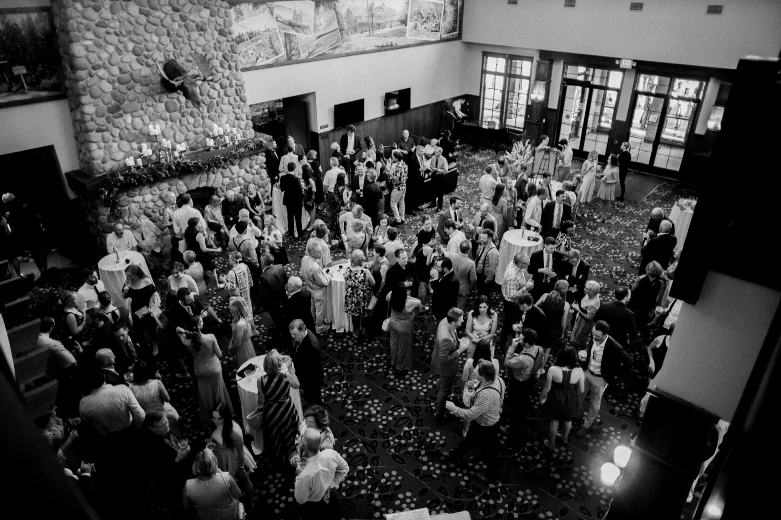 An aerial black-and-white view from a balcony shows over a hundred wedding guests mingling in the Grand View Lodge lobby beneath a massive stone fireplace with a mounted moose head. — Tim Larsen Photography, Brainerd Lakes MN