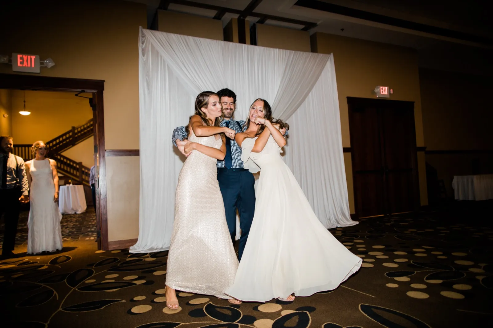 The bride and groom strike a playful pose with another bride in a white gown in front of a draped backdrop while a bridesmaid watches from a doorway at left. — Tim Larsen Photography, Brainerd Lakes MN