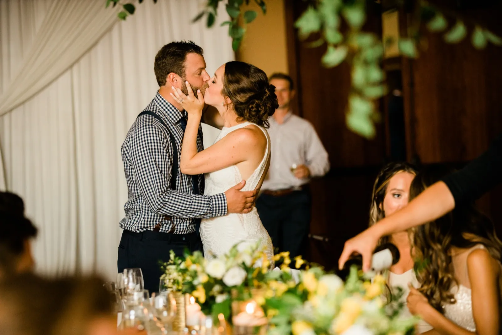 The bride and groom share a kiss behind the head table while guests look on, with eucalyptus garland hanging above and yellow and white florals on the table. — Tim Larsen Photography, Brainerd Lakes MN
