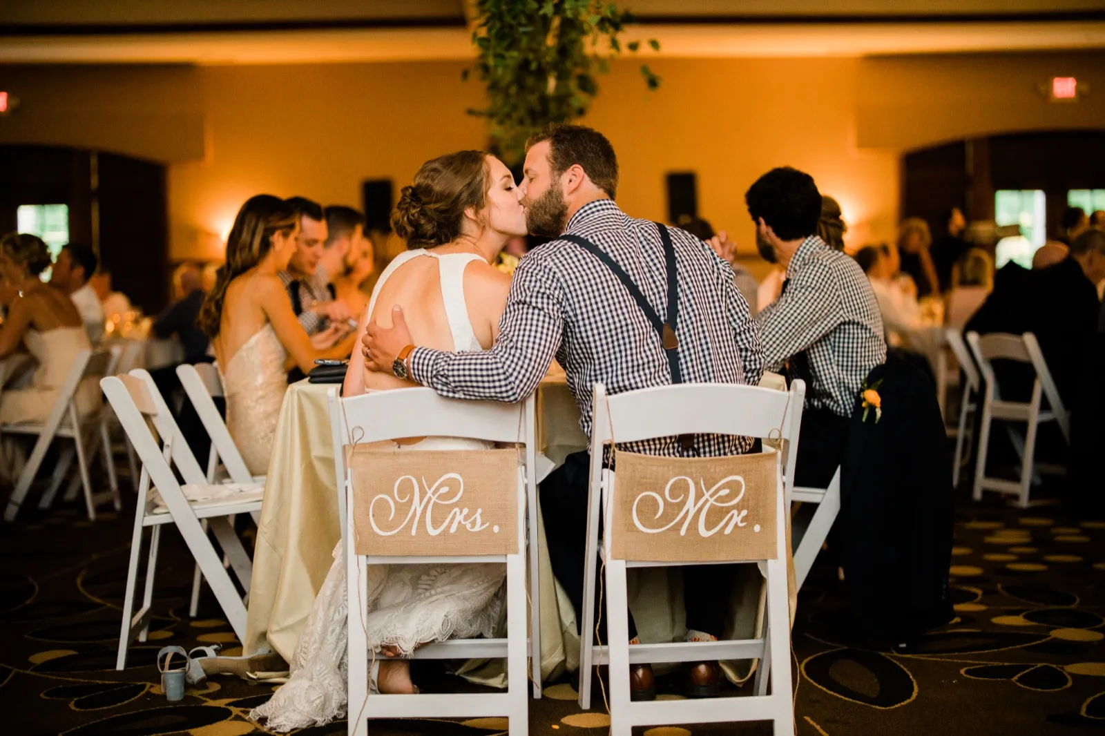 The bride and groom share a kiss at their sweetheart table, framed from behind by burlap chair signs reading Mr. and Mrs. in a warmly lit reception ballroom. — Tim Larsen Photography, Brainerd Lakes MN