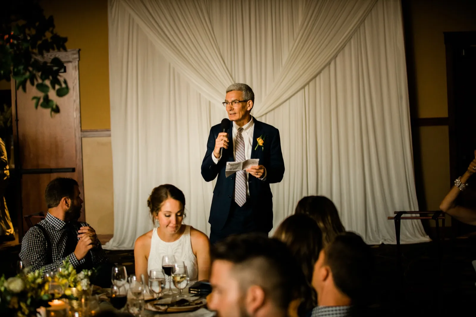 A man in a navy suit holds a microphone and notes while giving a speech against a white draped backdrop, with the bride listening at the foreground table. — Tim Larsen Photography, Brainerd Lakes MN