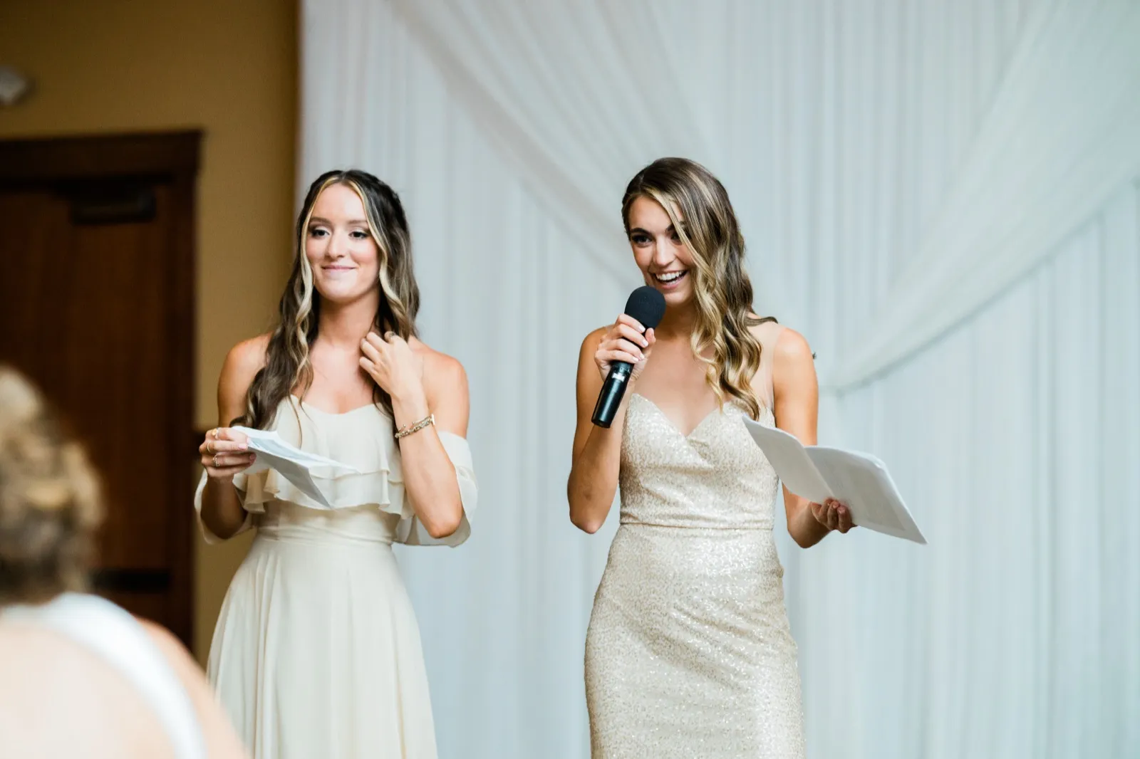 Two bridesmaids stand together giving a toast, one holding a microphone and laughing brightly while the other smiles holding her speech notes. — Tim Larsen Photography, Brainerd Lakes MN