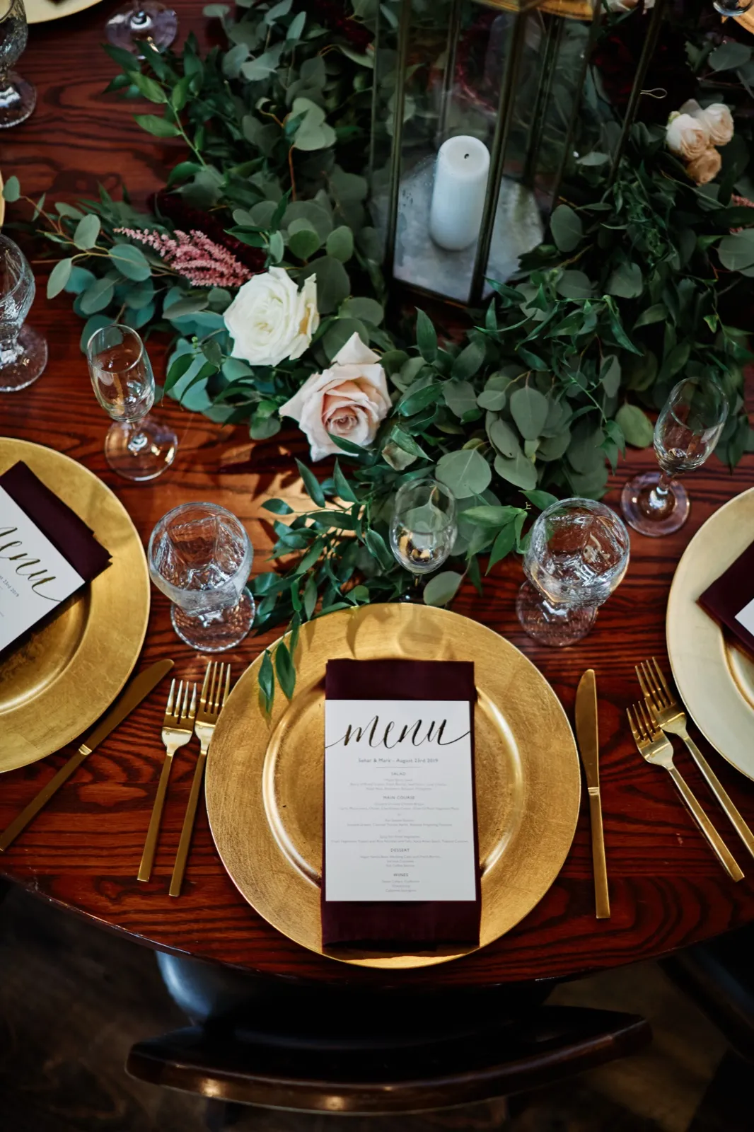 An overhead view of a reception place setting with a gold charger, burgundy menu napkin, gold flatware, and a greenery and rose garland centerpiece with a lit pillar candle. — Tim Larsen Photography, Brainerd Lakes MN