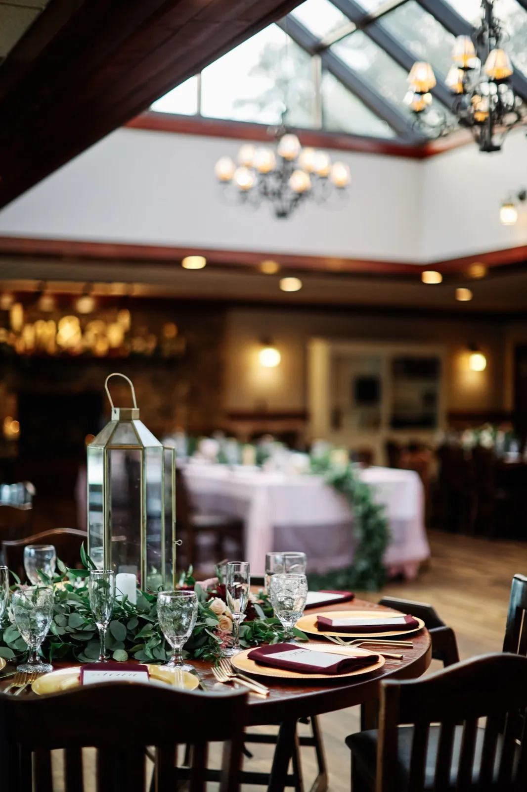 A reception table with a tall metal lantern, greenery garland, and burgundy napkins sits beneath the skylit beams and chandeliers of the Grand View Lodge dining room. — Tim Larsen Photography, Brainerd Lakes MN