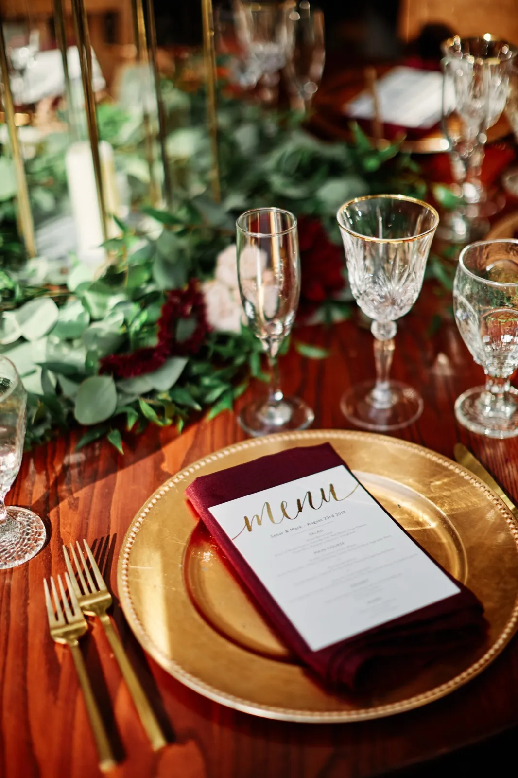 A gold charger plate holds a burgundy napkin and a gold-script menu card dated August 23rd 2019, surrounded by gold-rimmed crystal glassware and a eucalyptus and rose garland. — Tim Larsen Photography, Brainerd Lakes MN
